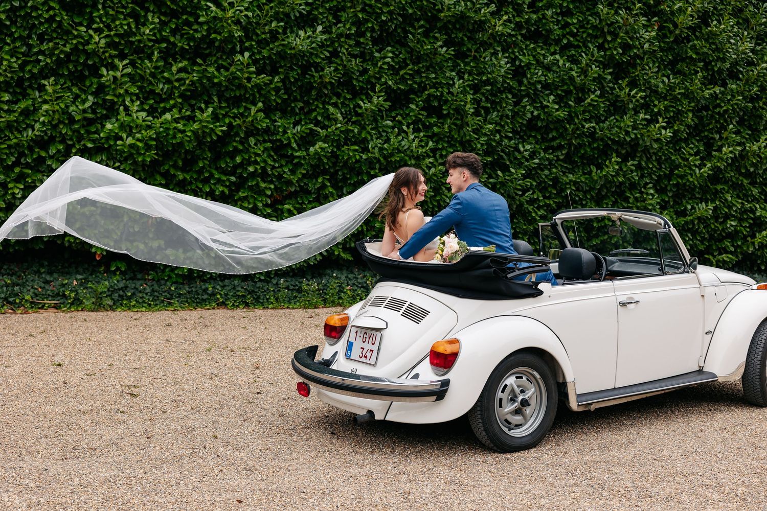 Bride and groom sit together in white vintage VW Beetle convertible with wedding veil flowing in breeze.