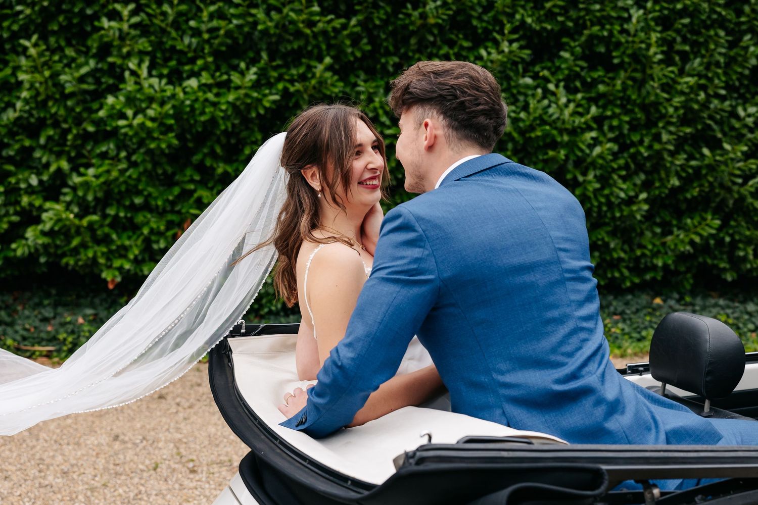 Bride and groom sitting together in a classic convertible car smiling at each other.