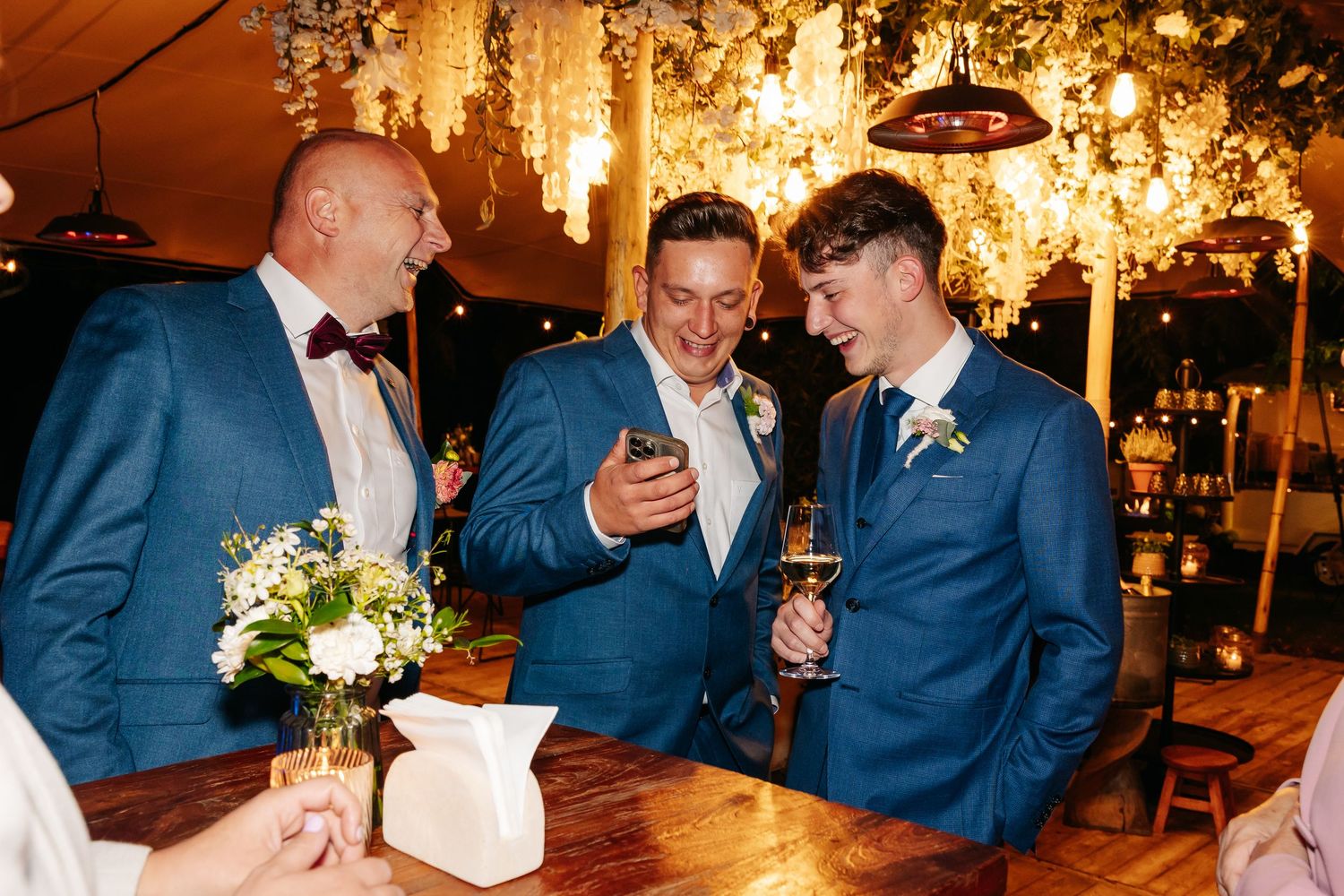 Three men in blue suits laughing and sharing phone screen at wedding reception bar.