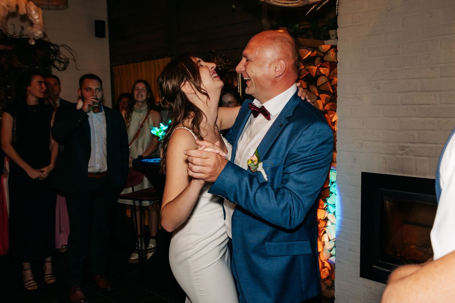 Bride laughing joyfully while dancing with a man in a blue suit at wedding.