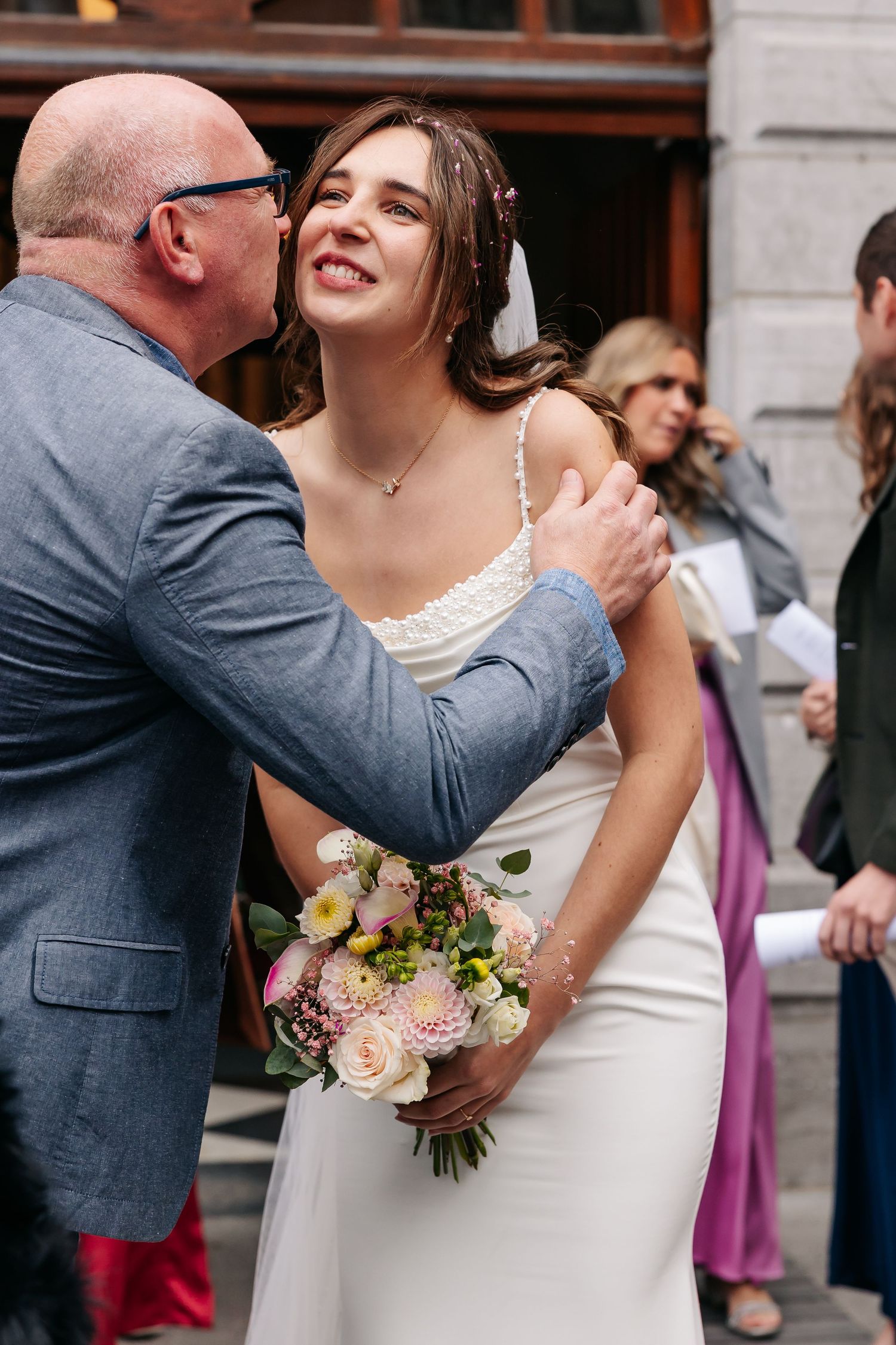 Bride in white gown holding pastel bouquet receives a warm kiss on the cheek from an older male guest.