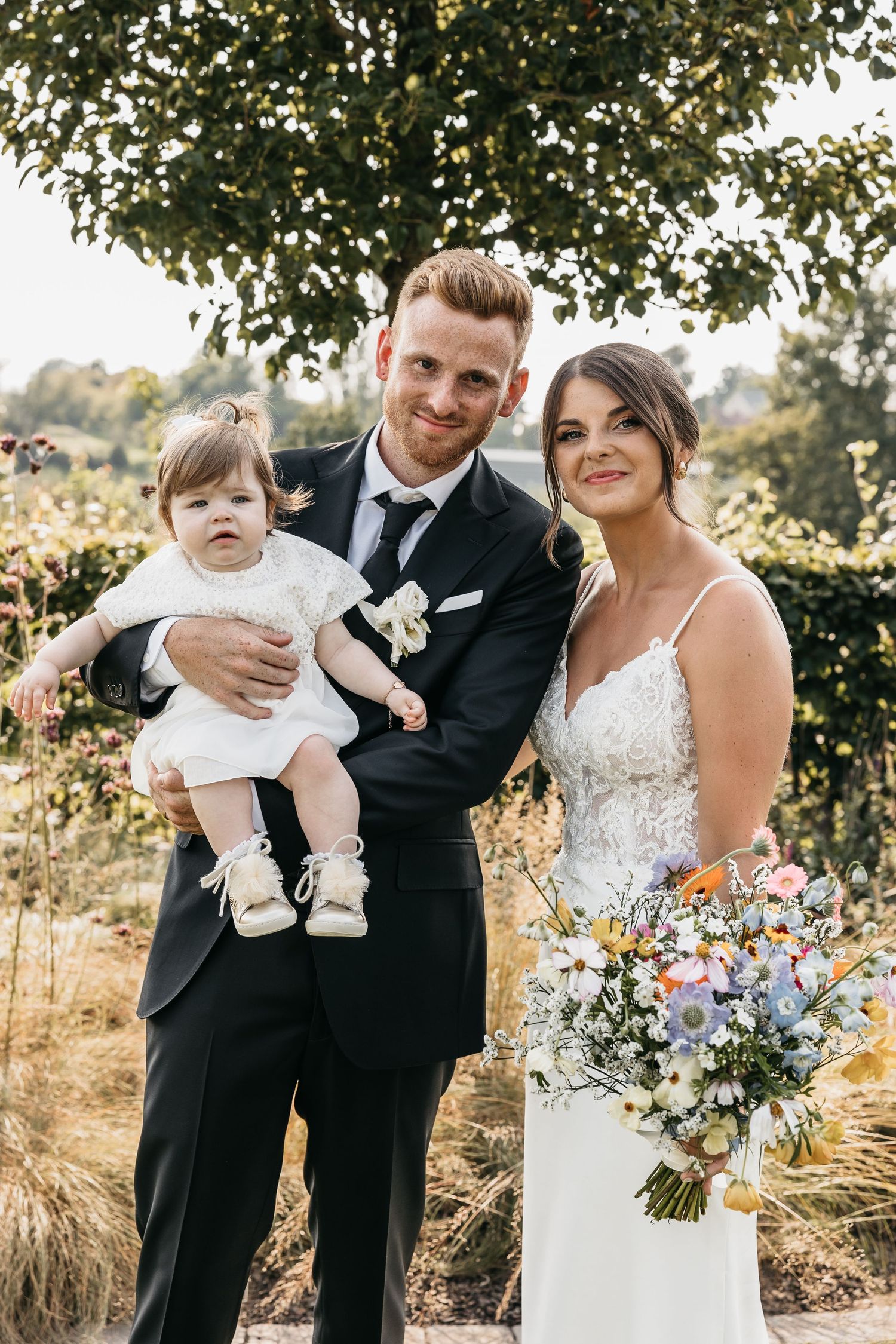 Groom in black suit holds baby girl in white dress beside bride in lace gown with wildflower bouquet outdoors.