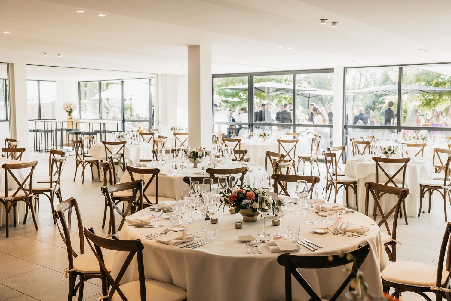 Bright wedding reception hall with round tables, white linens, wooden chairs, and large windows.
