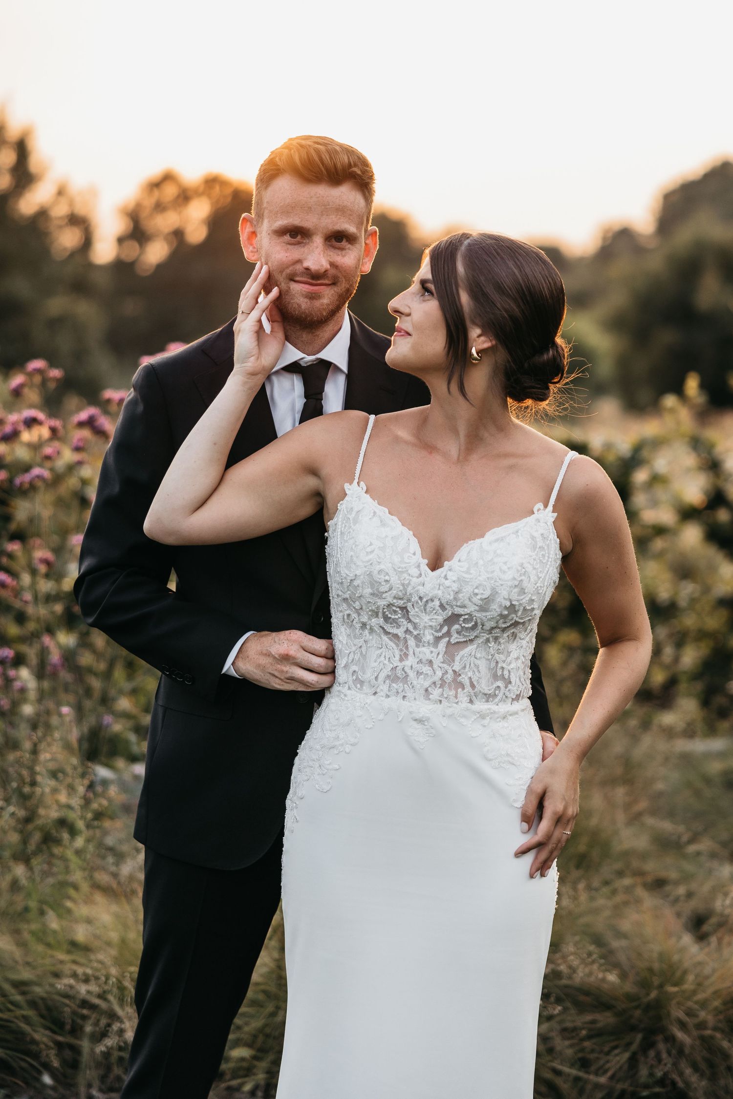 Bride in white lace gown tenderly touches groom's face as they smile at each other in a sunlit garden.