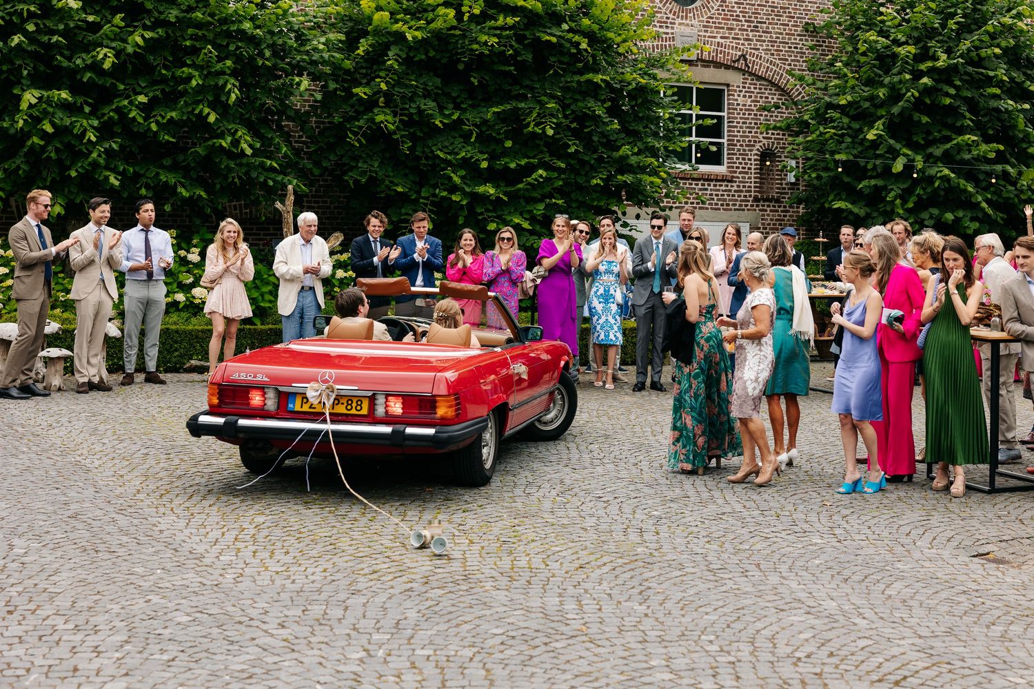 Bride and groom departing in red vintage convertible as wedding guests celebrate on courtyard.