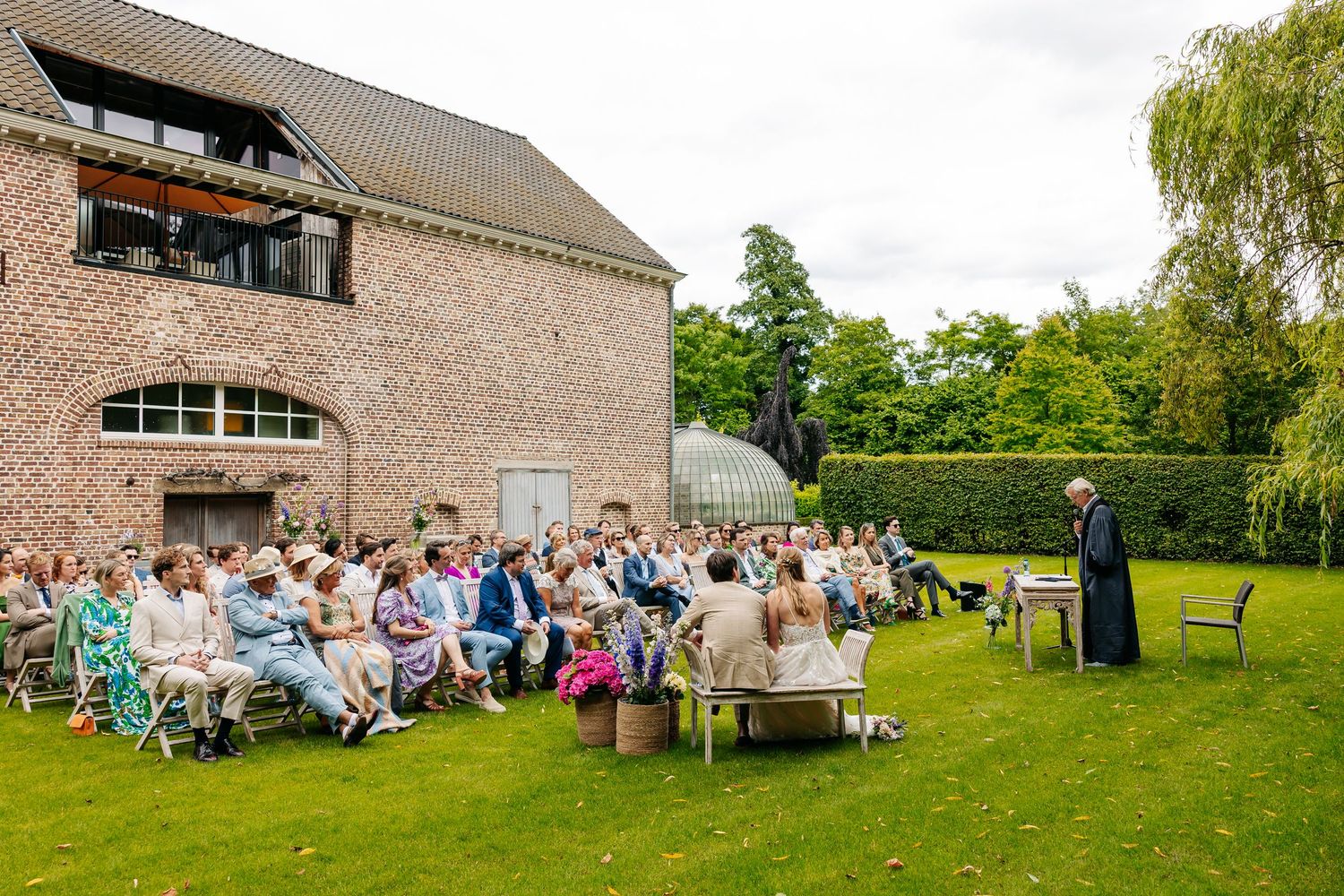 Wedding guests seated outdoors on lawn chairs during a garden ceremony beside a rustic brick building.