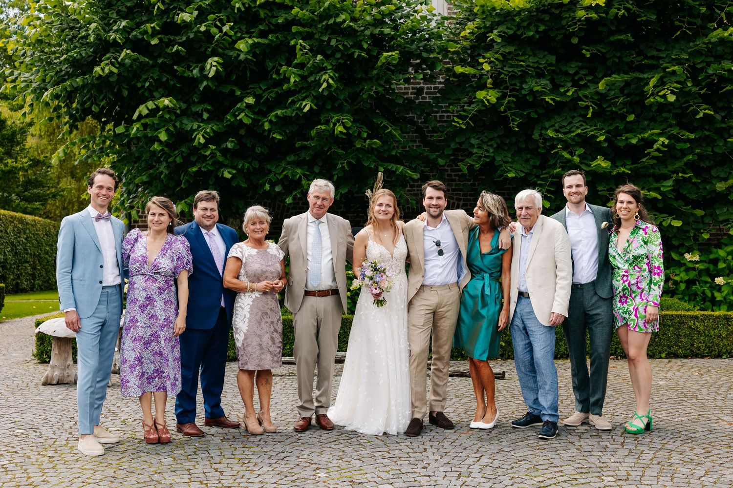 Wedding party group portrait of ten guests and couple posing on cobblestone path with green hedges.
