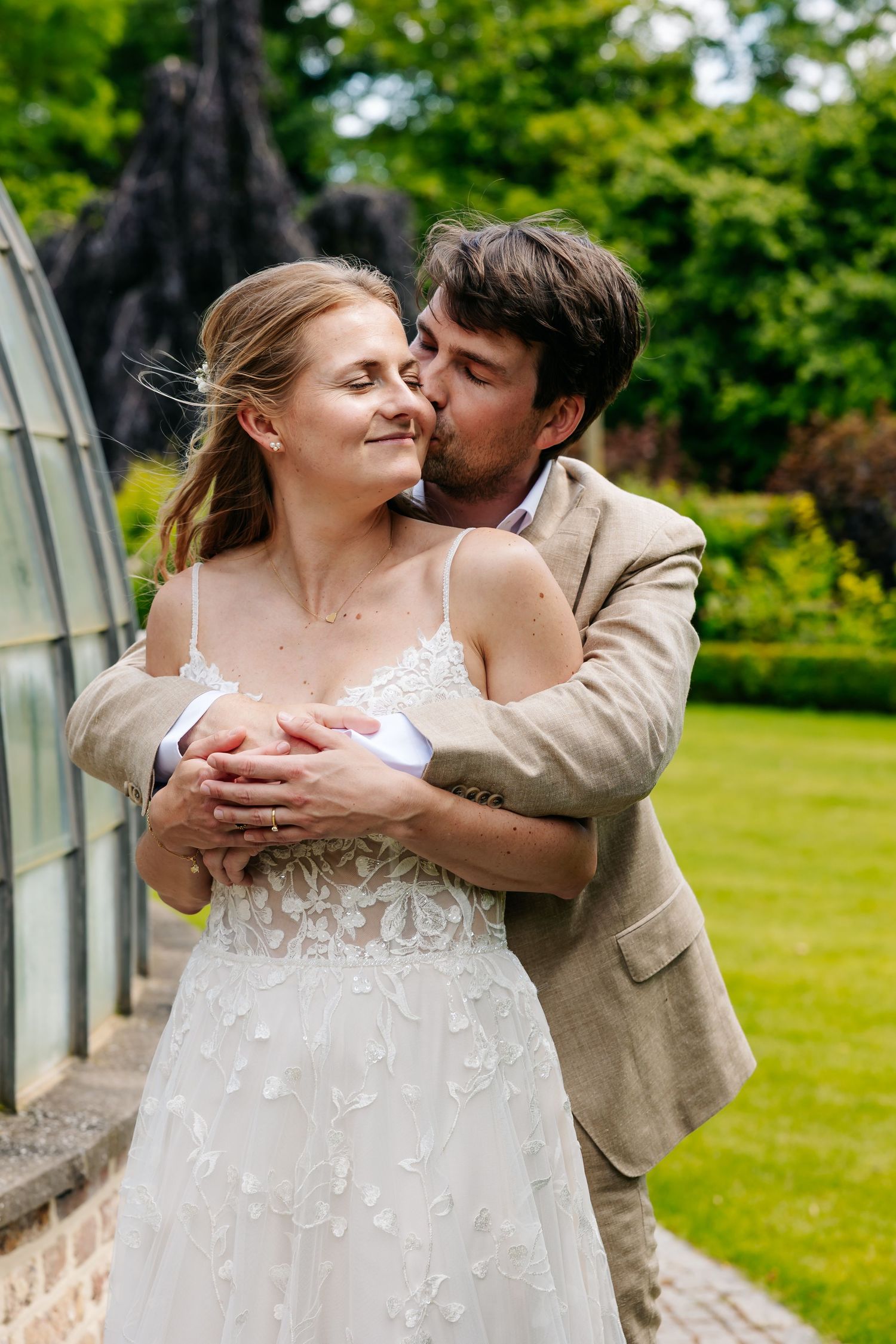 Groom tenderly kisses bride on the cheek while embracing her near a glass greenhouse in lush gardens.