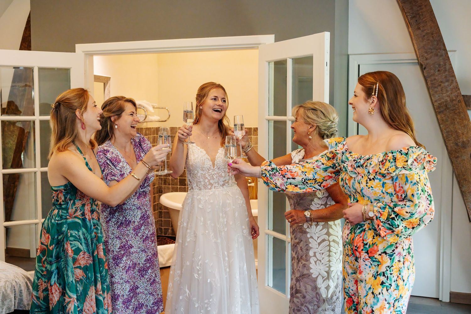 Bride in lace wedding gown toasting champagne with four bridesmaids in colorful floral dresses during wedding preparations.
