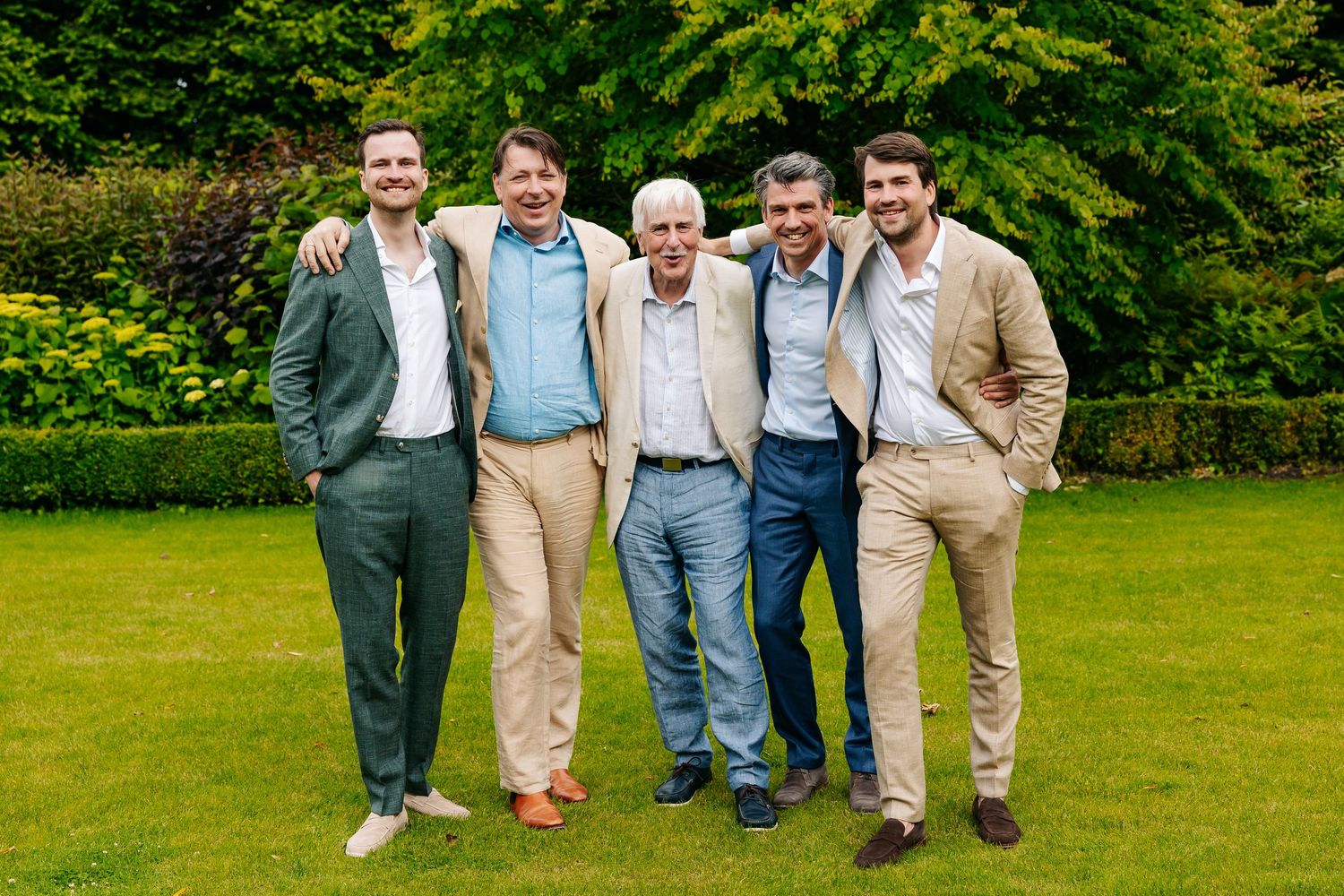 Five men in suits smile together on a green lawn at a summer wedding celebration.