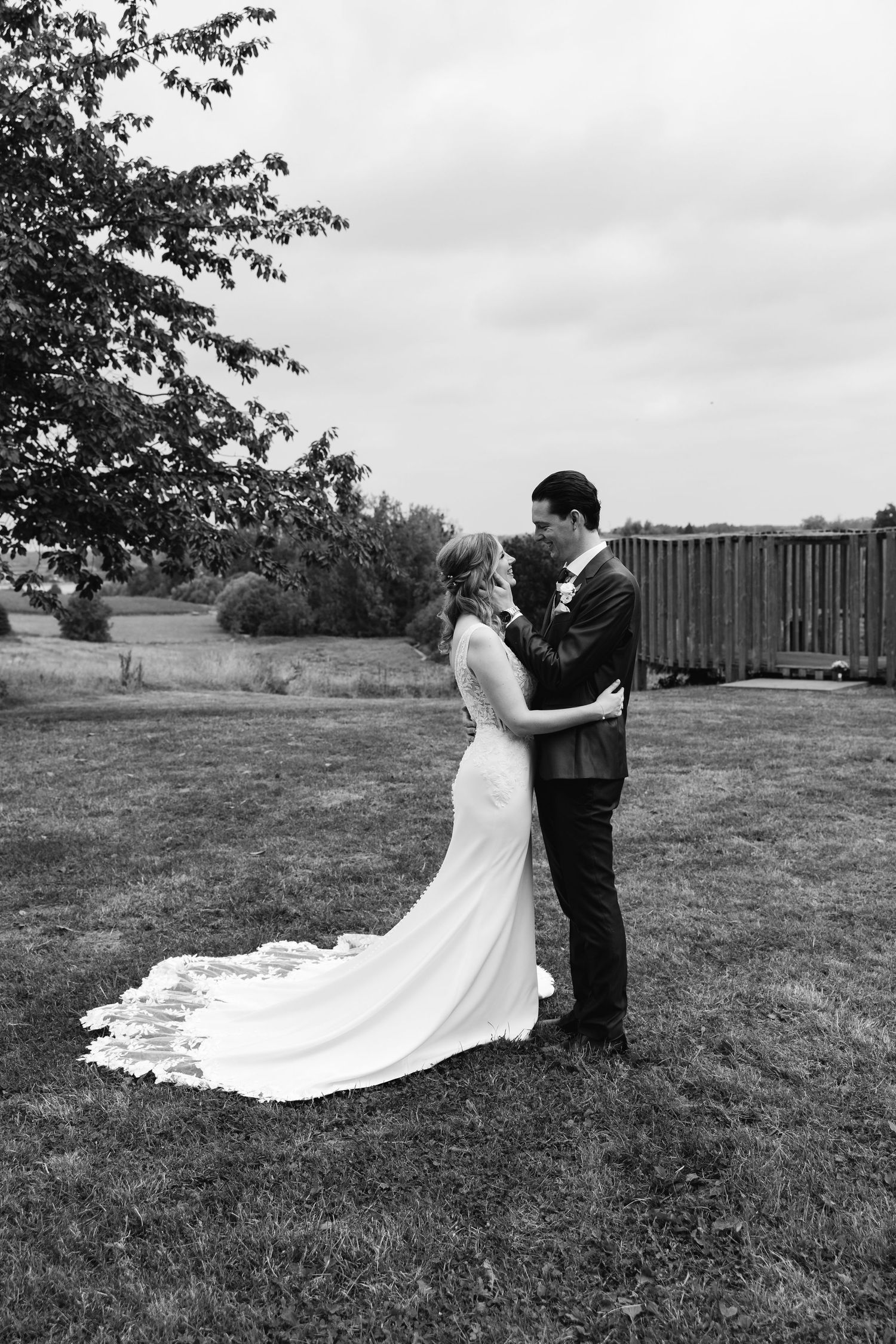 Black and white photo of bride and groom embracing on a lawn with trees and wooden fence behind them.