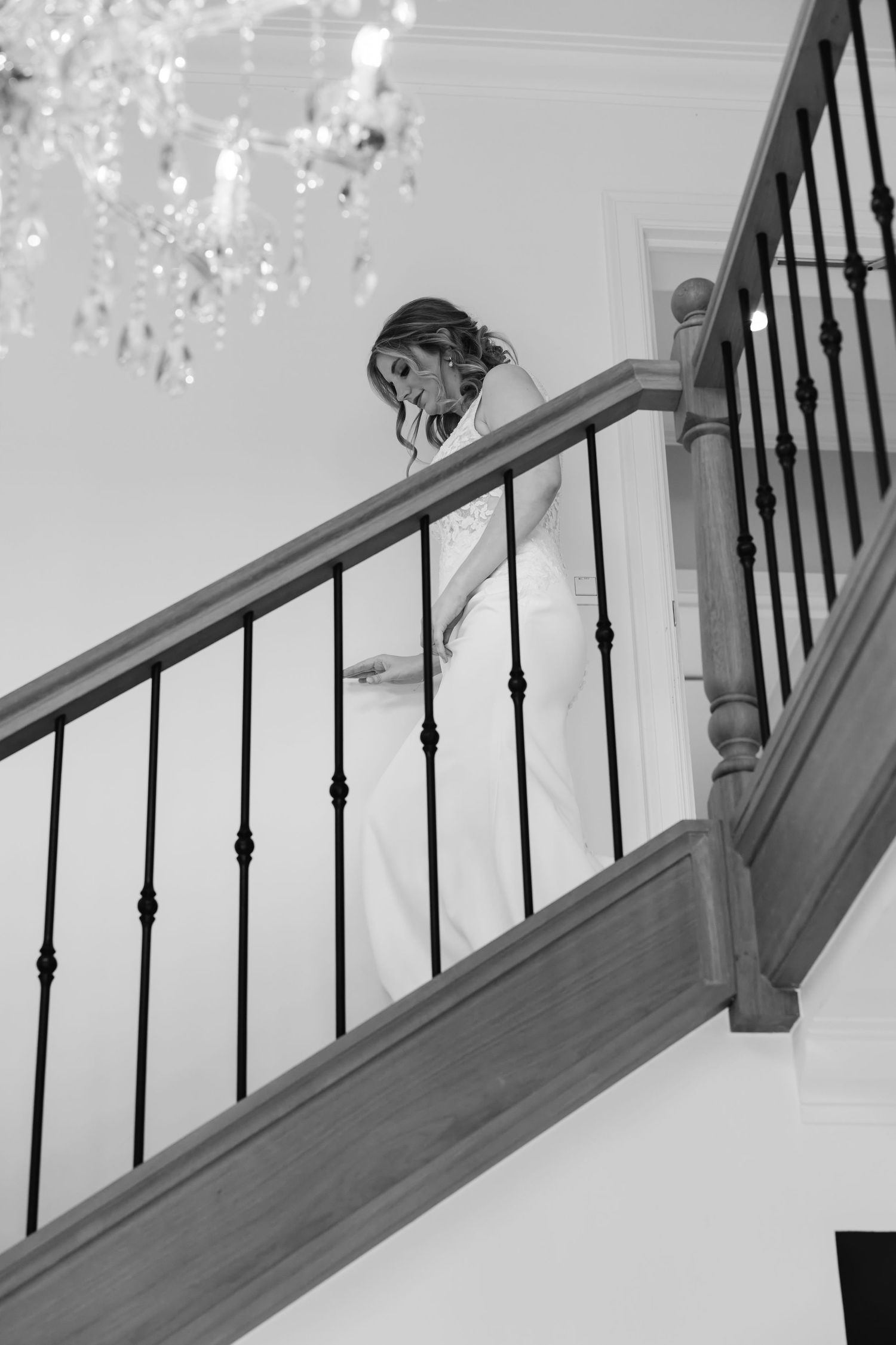 Elegant bride in white wedding gown descends a wooden staircase beneath a crystal chandelier, black and white.