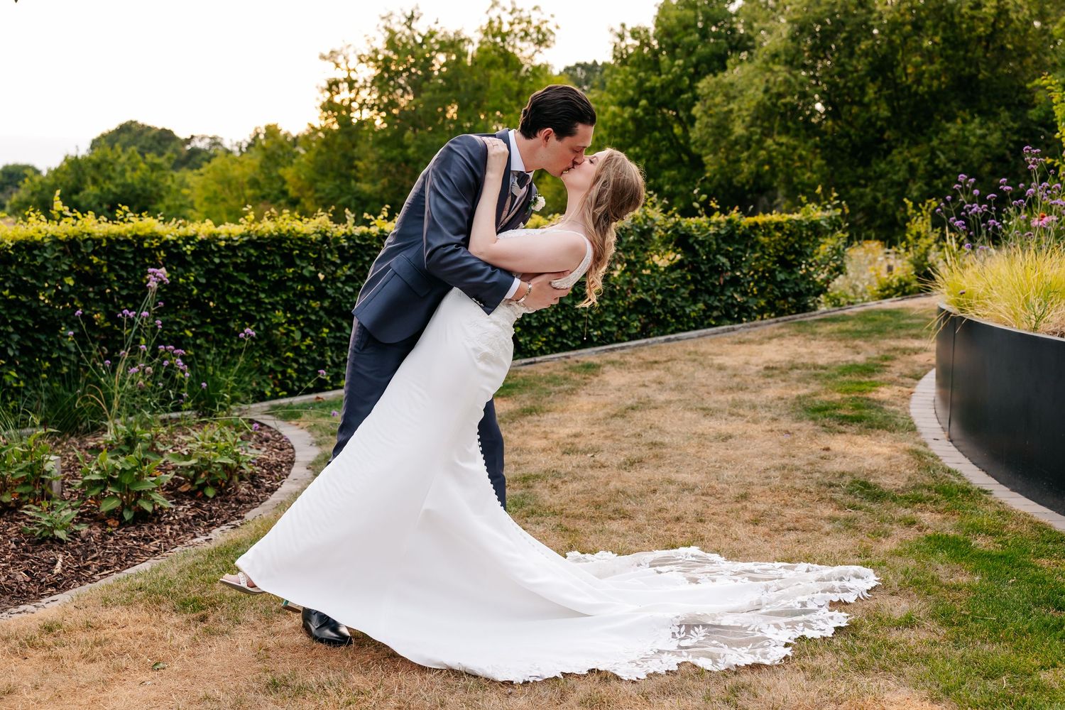 Groom dips bride in a white gown for a romantic kiss in a garden with a neatly trimmed hedge.