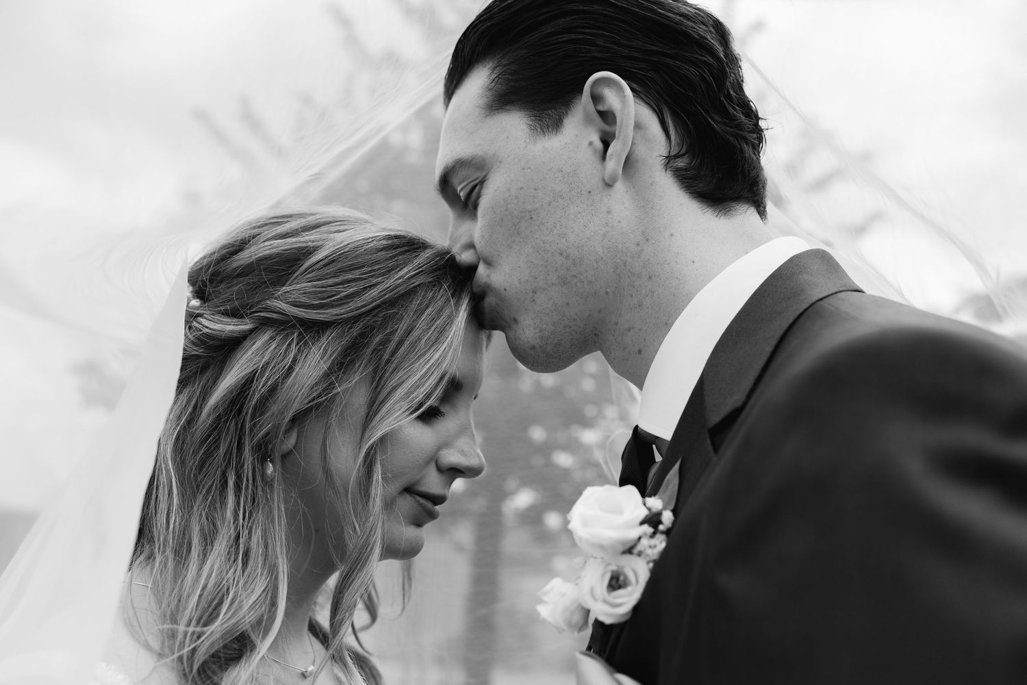 Black and white close-up of groom kissing brides forehead tenderly beneath a flowing veil.