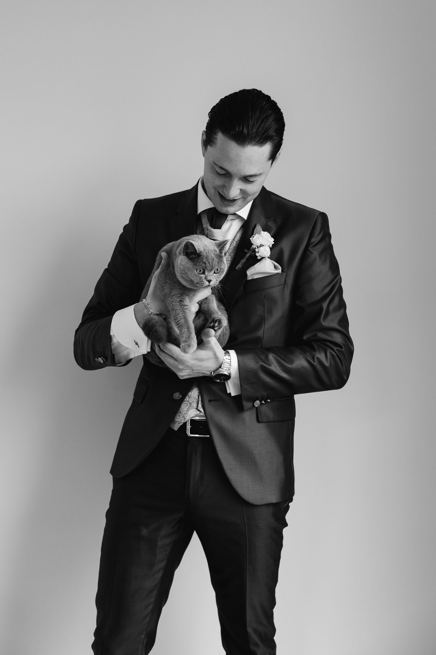 Black and white portrait of a groom in navy suit holding a gray British Shorthair cat, smiling.