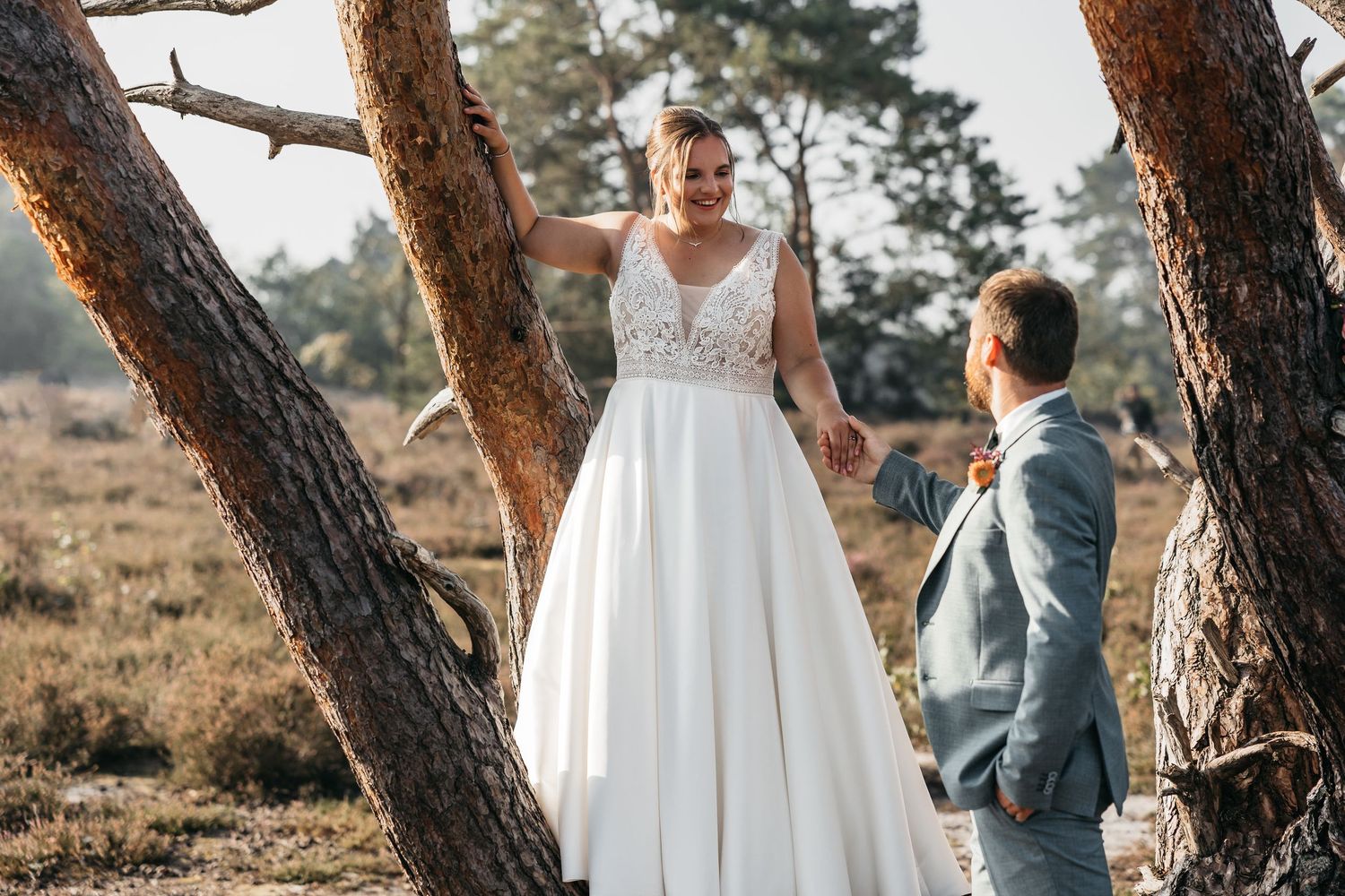 Smiling bride in white lace gown holds groom's hand near pine trees in a sunlit heathland landscape.