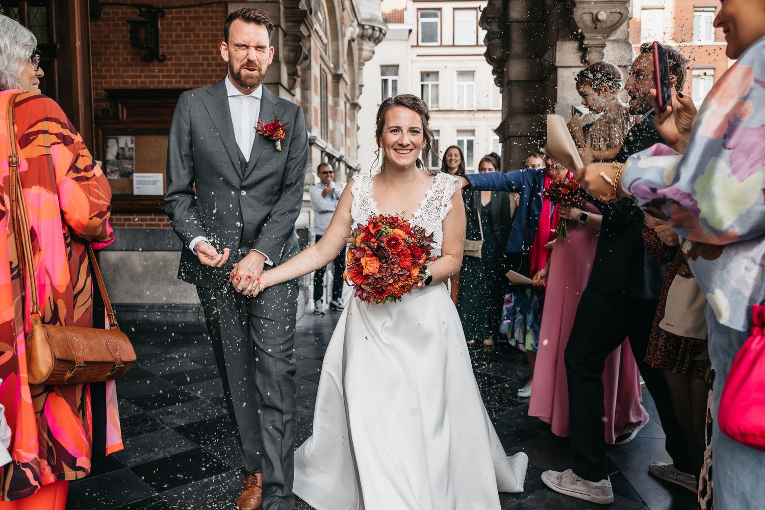 Happy bride holding autumn bouquet walks with groom through confetti shower outside wedding venue.