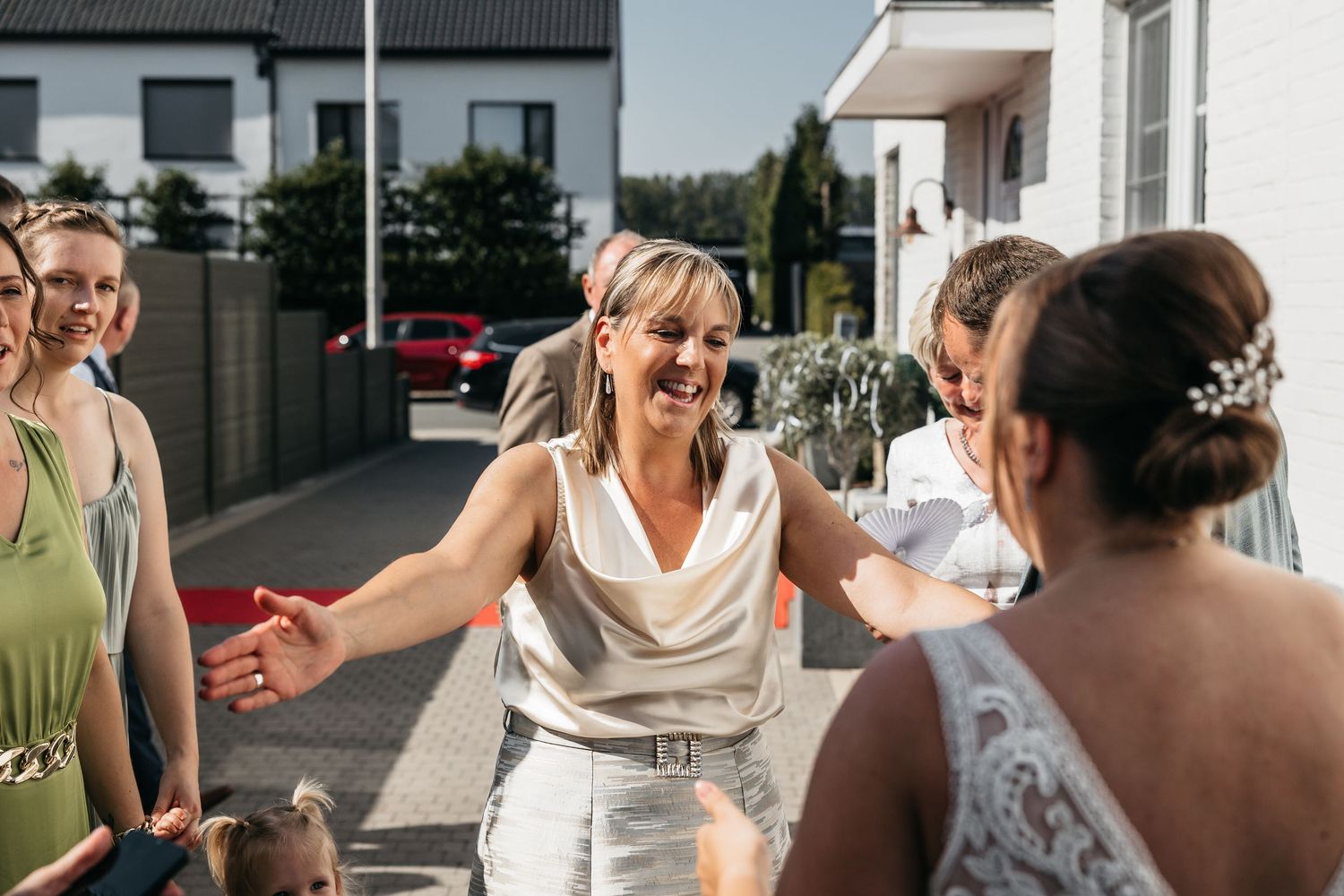 Joyful woman in silver dress laughs and opens her arms to greet the bride at a sunny wedding.