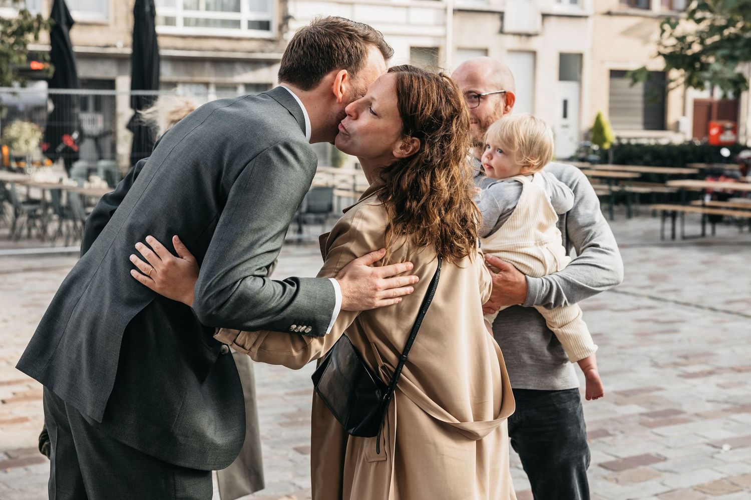 Man in gray suit sharing a warm kiss with woman holding a baby on a city plaza.