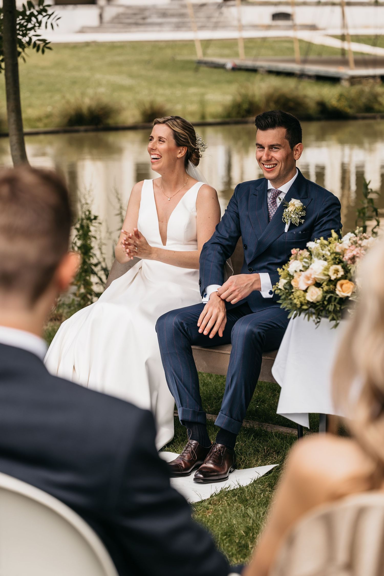 Laughing bride and groom sit together during outdoor wedding ceremony beside a serene pond.