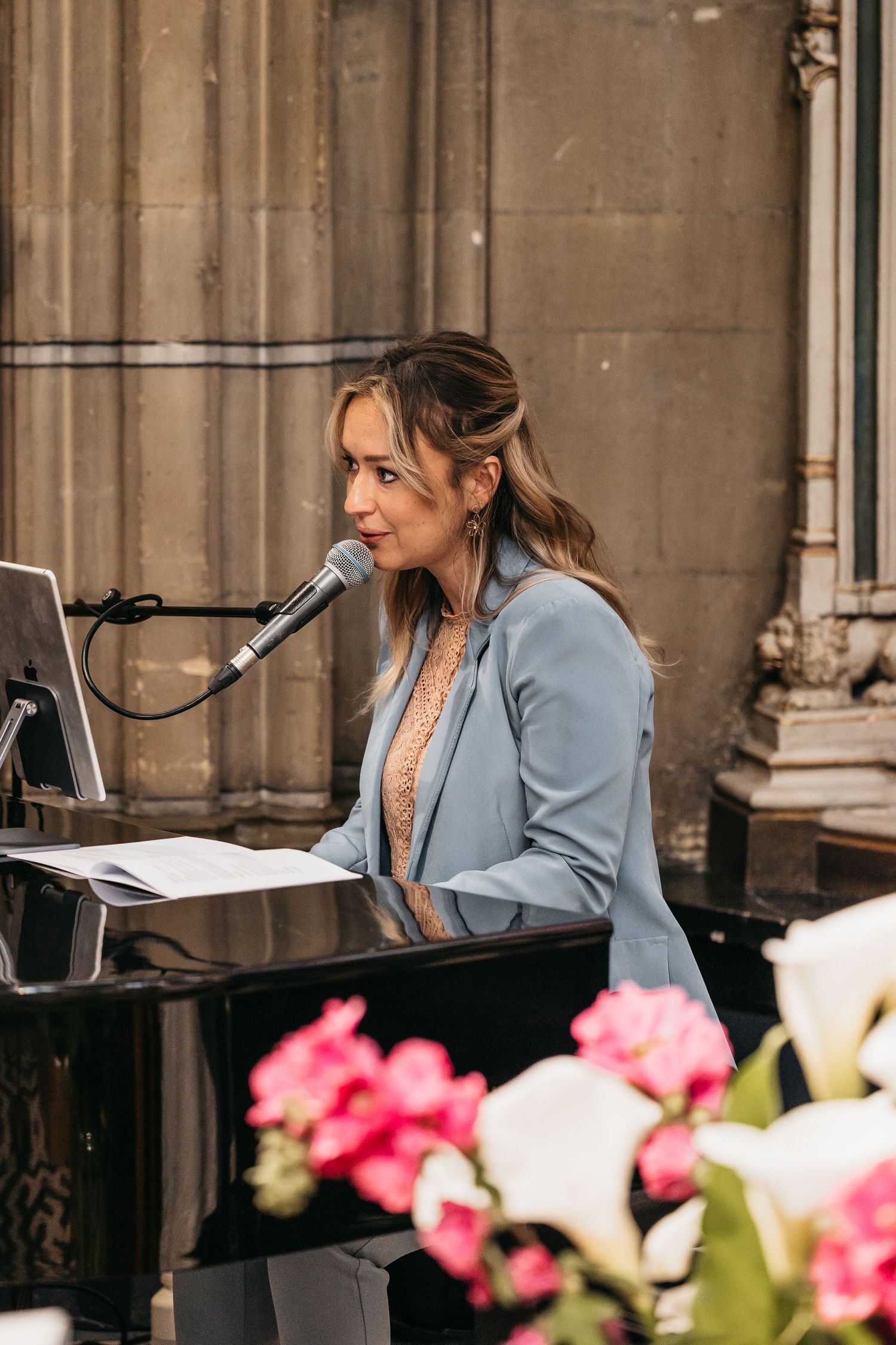 Woman in blue blazer performing at piano with microphone during elegant church wedding ceremony.
