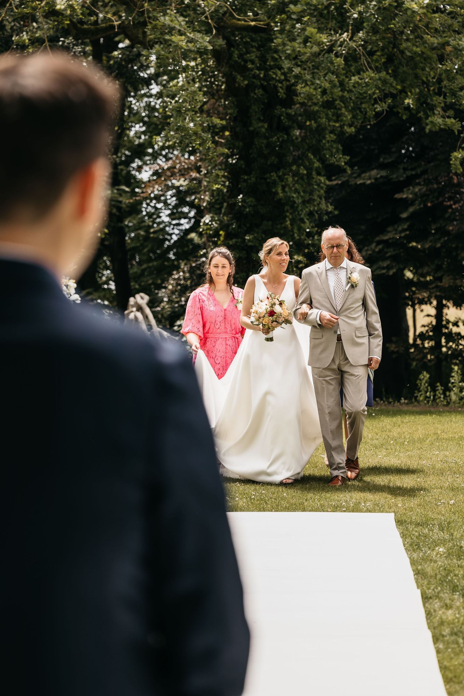Bride walks down outdoor aisle escorted by father and bridesmaid toward waiting groom.