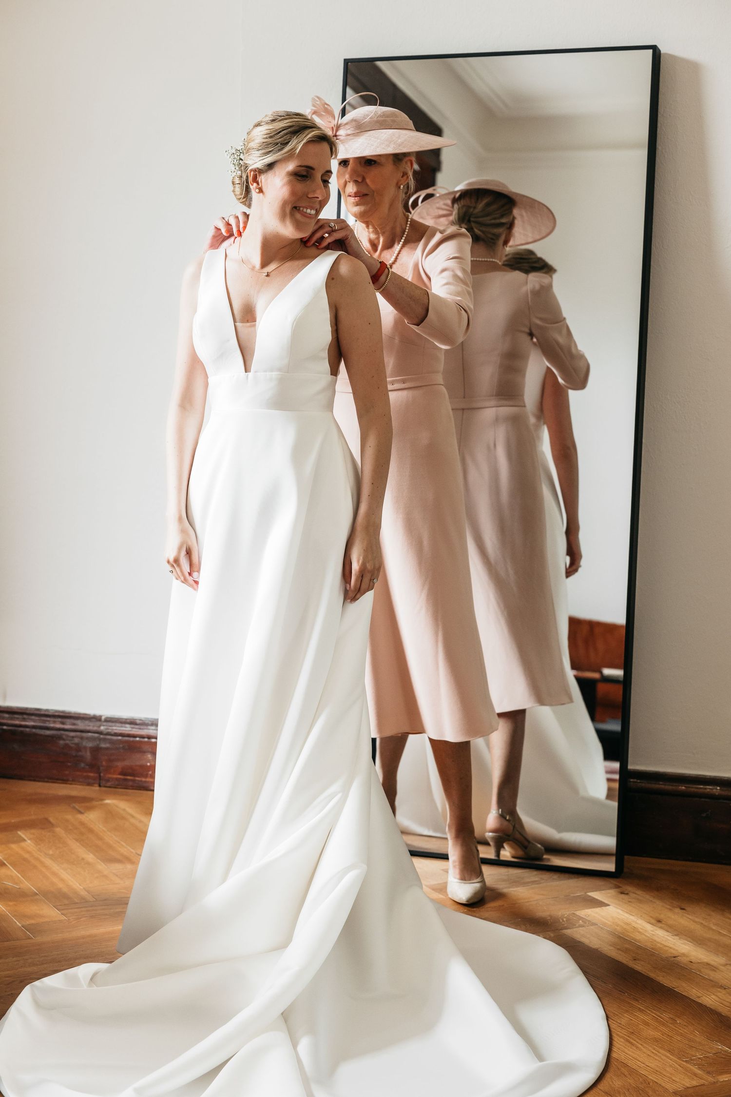 Bride in white gown stands with older woman in pink dress and hat before mirror.