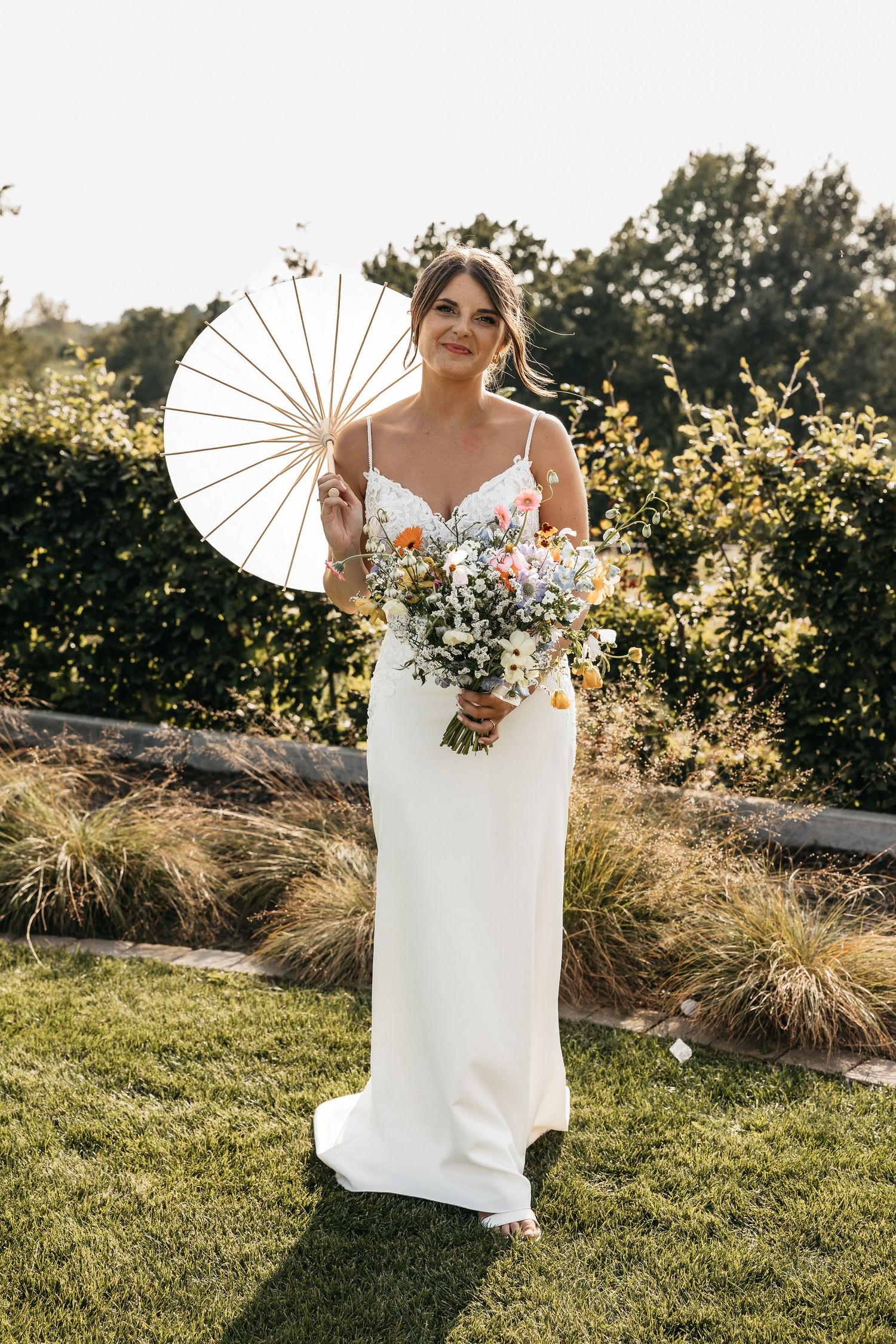 Beautiful bride in white spaghetti strap gown holding parasol and wildflower bouquet on a sunny lawn.