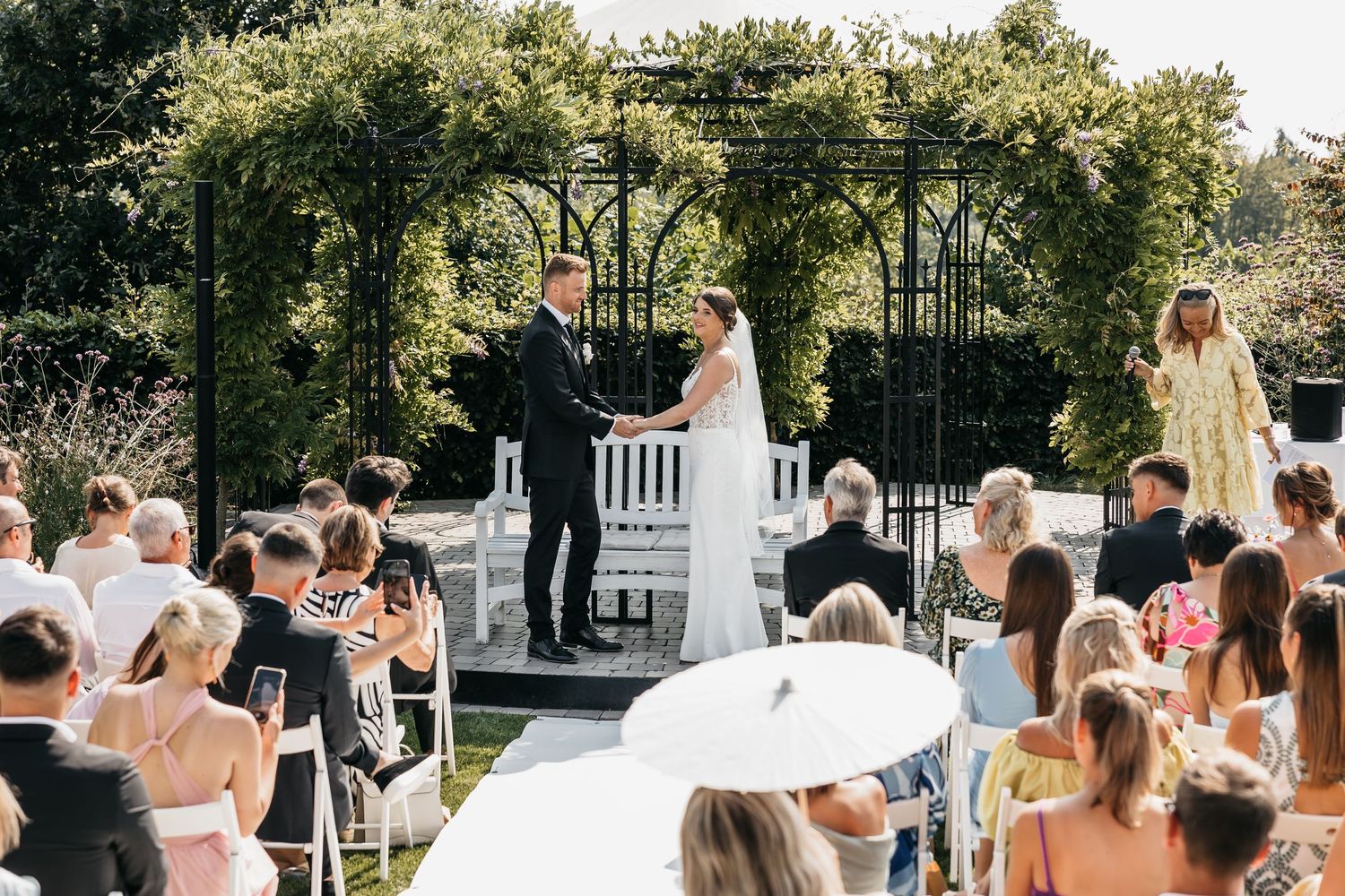 Bride and groom hold hands at outdoor wedding altar beneath black iron pergola arch.