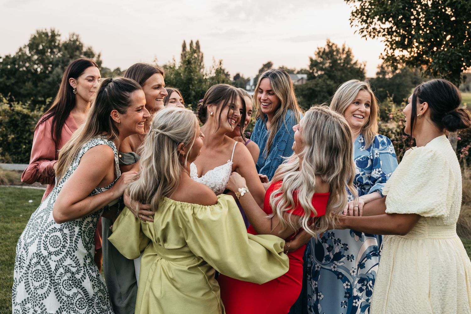 Bride surrounded by laughing female guests sharing joyful moment at outdoor wedding reception.