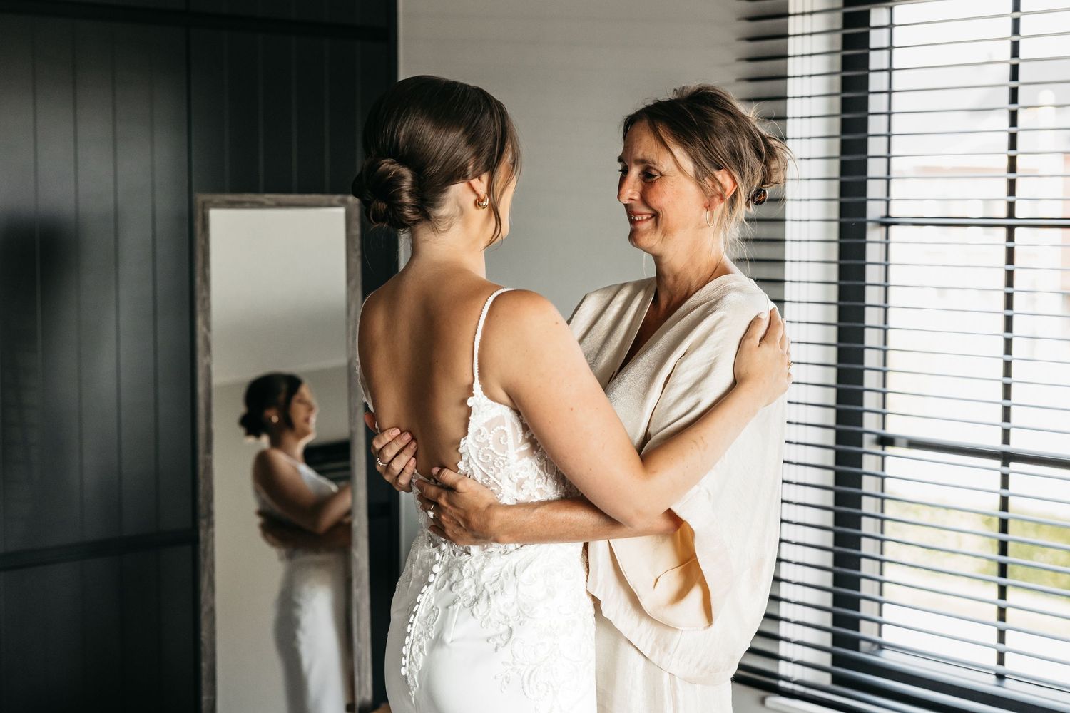 Bride in white lace wedding dress embracing woman in beige robe near venetian blinds.