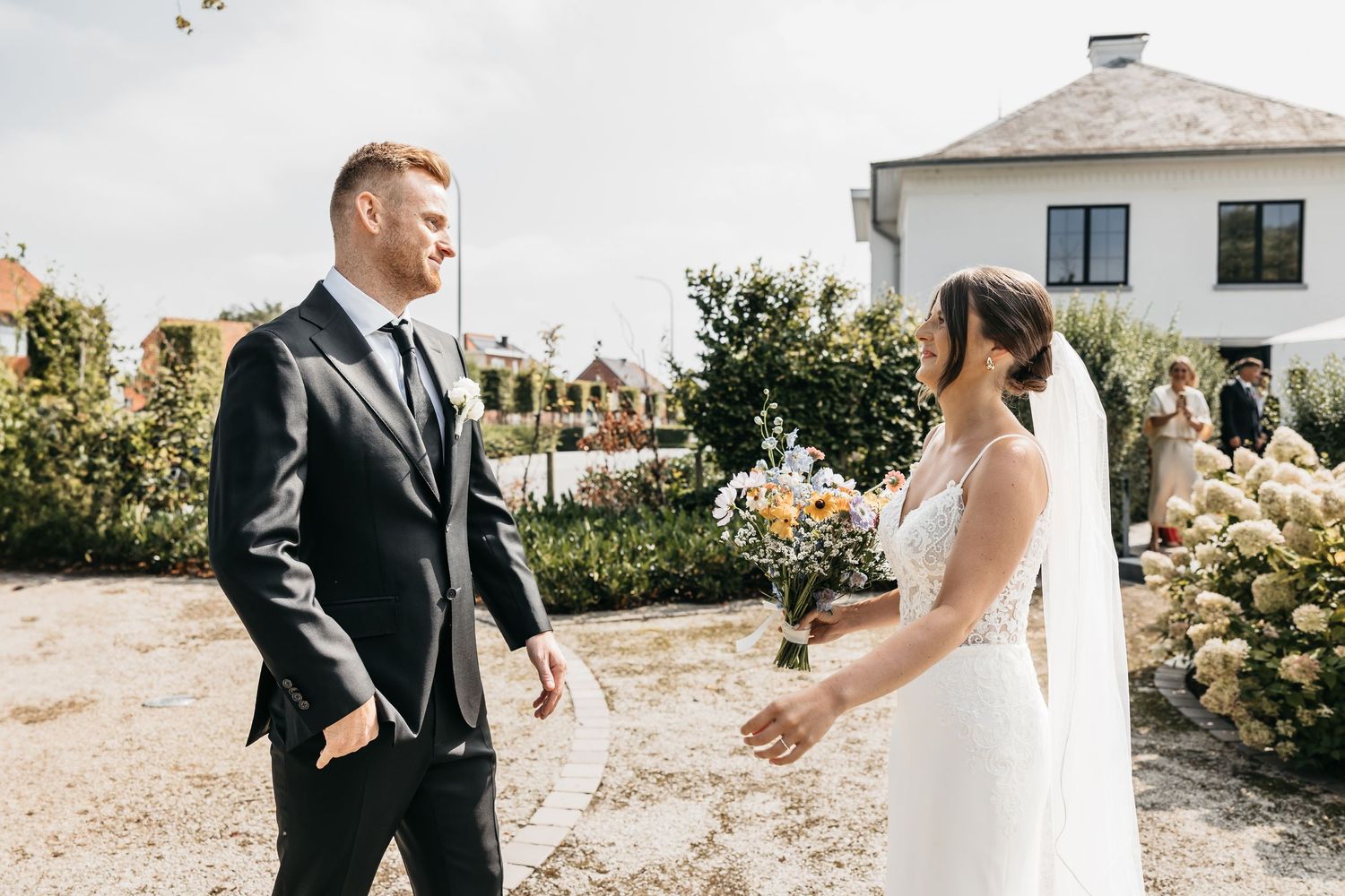 Bride holding colorful bouquet faces groom during first look in sunlit garden setting.