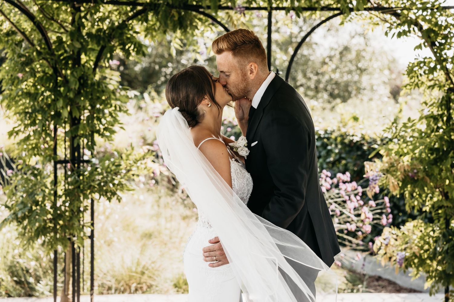 Bride and groom kissing under lush green garden arch in wedding attire.