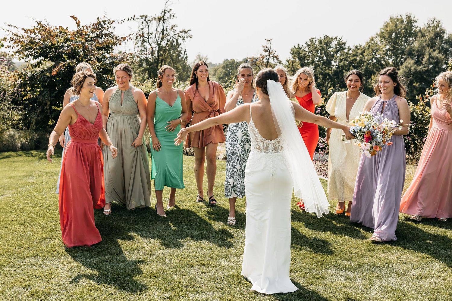 Bride tosses wedding bouquet to eager bridesmaids wearing colorful mismatched gowns on a sunny outdoor lawn.