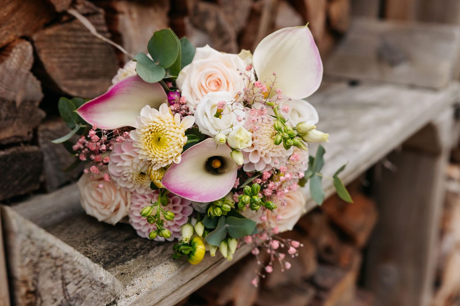 Elegant bridal bouquet with calla lilies, roses, and dahlias resting on a rustic wooden bench.