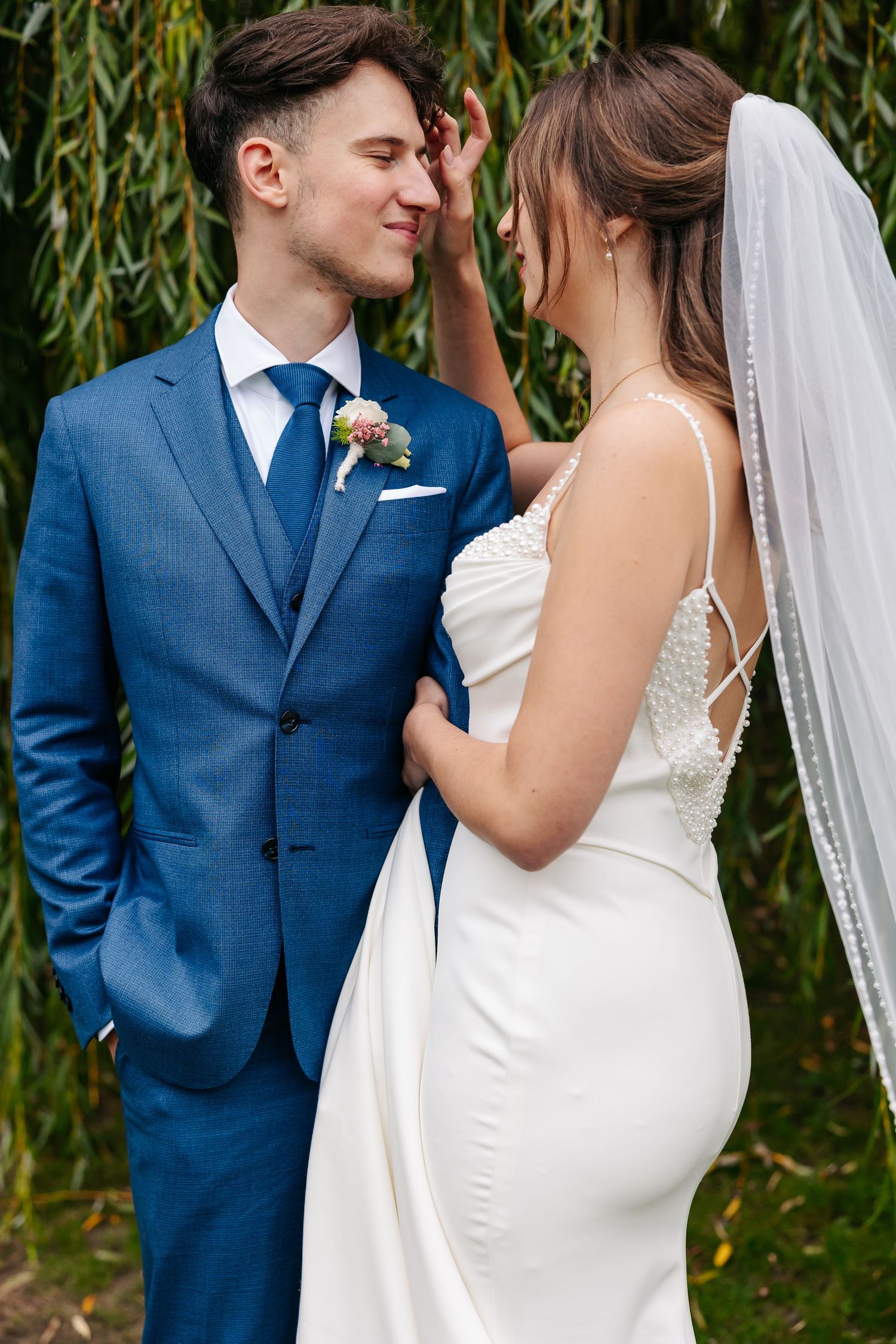 Bride and groom share intimate moment under willow tree, groom in blue suit smiling at veiled bride.