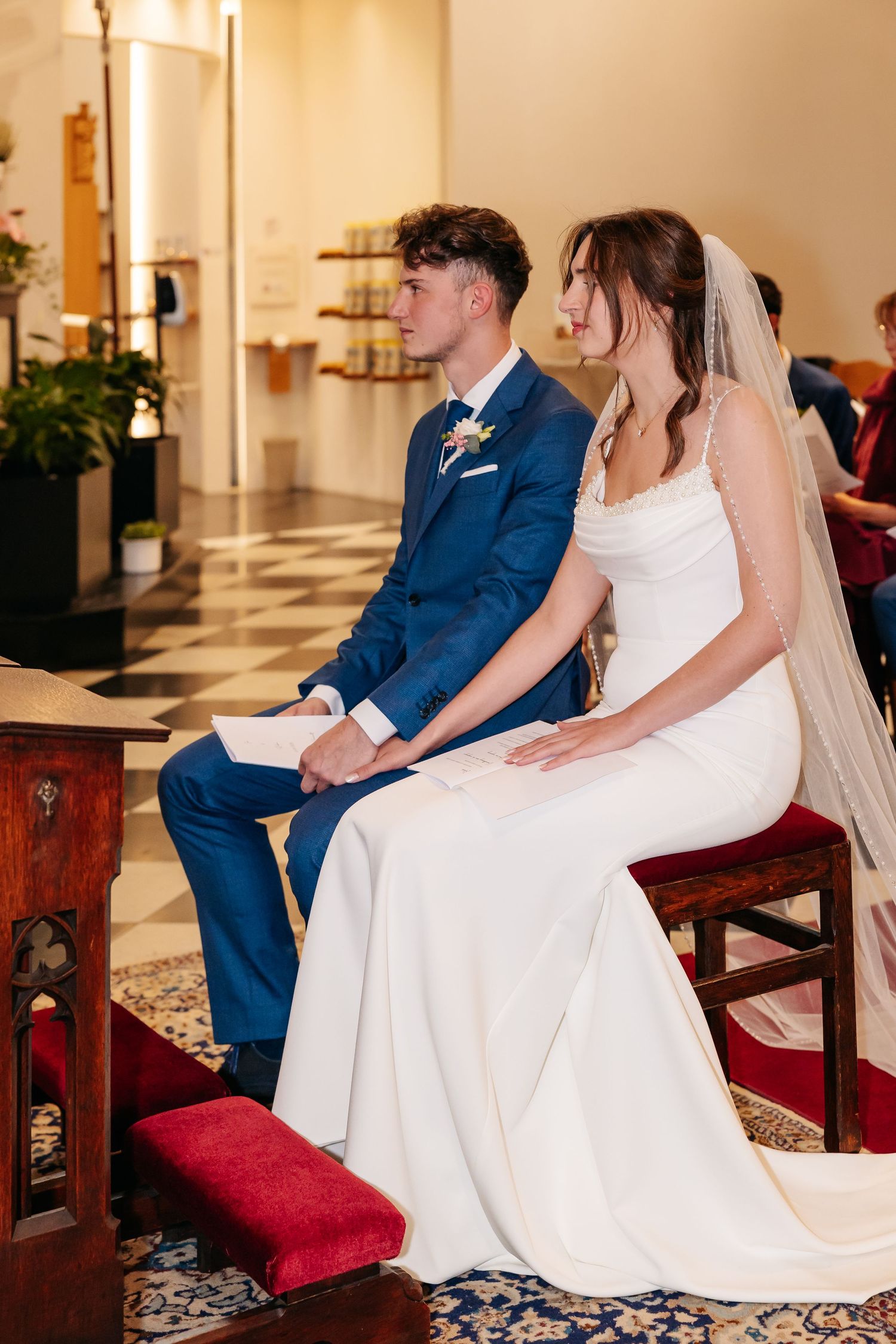 Bride in white gown and groom in blue suit sit together during church wedding ceremony.