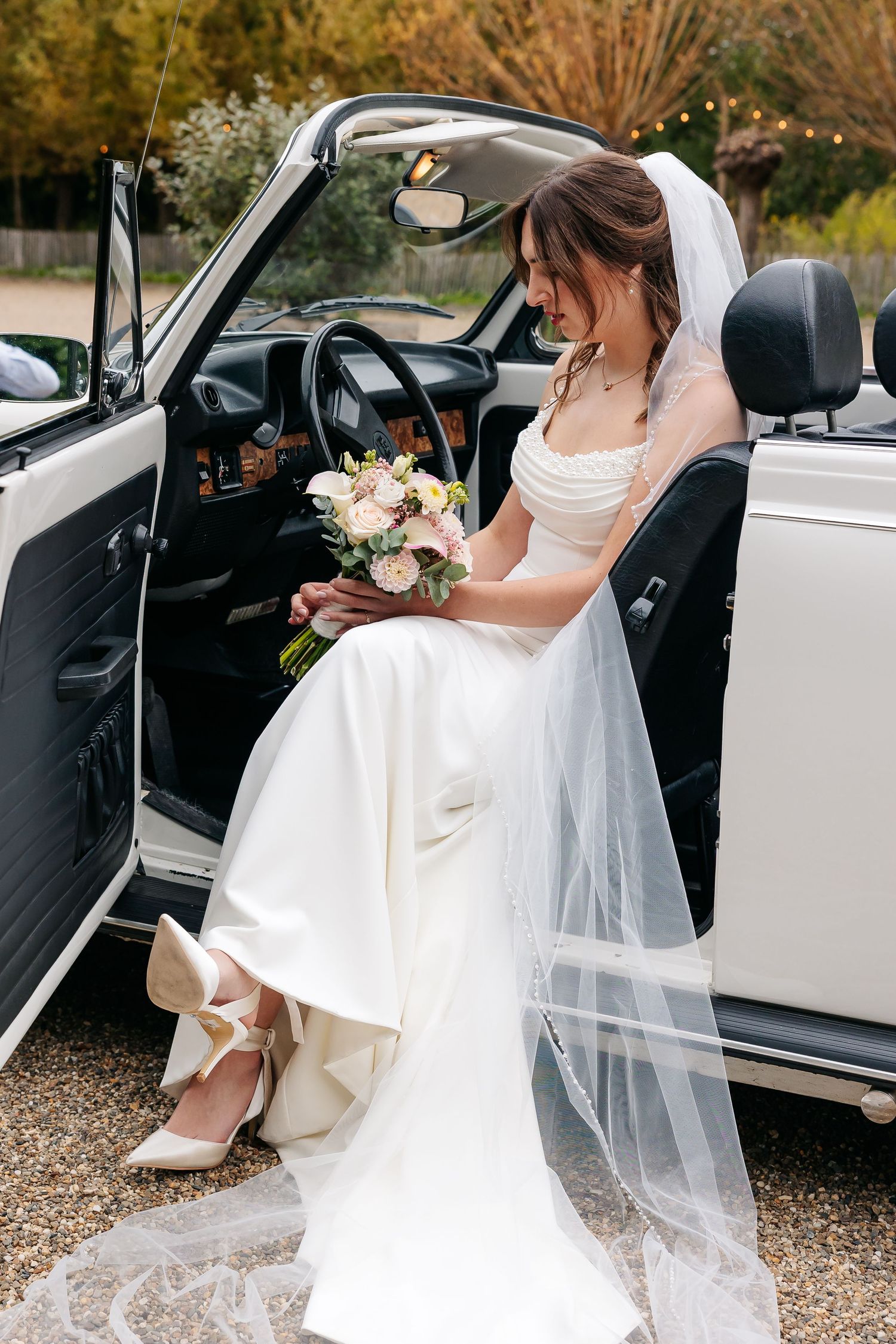 Elegant bride in white gown holding a floral bouquet seated in a classic white vintage convertible wedding car.
