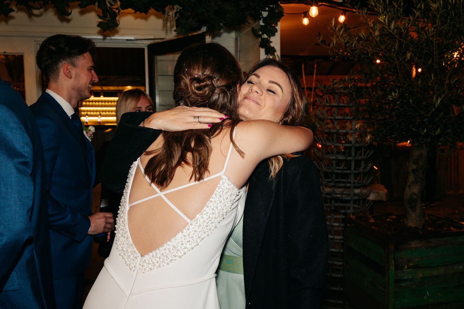 Bride in backless white gown sharing a warm hug with a guest at wedding reception.