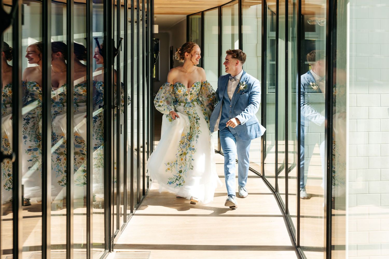 Bride in floral dress and groom in blue suit walking through mirrored glass corridor at wedding.