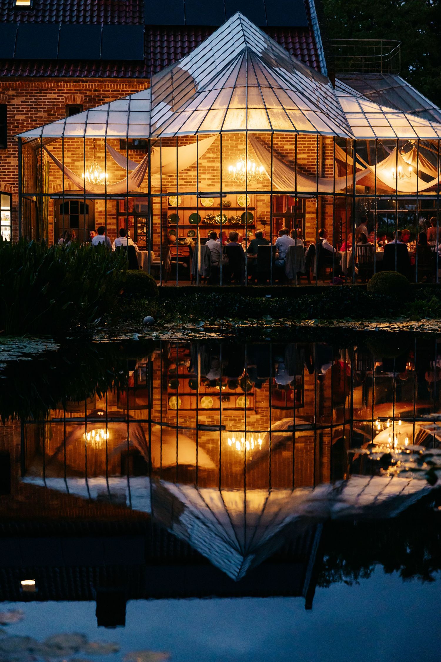 Stunning glass conservatory wedding venue with warm lights reflected in a serene pond at night.