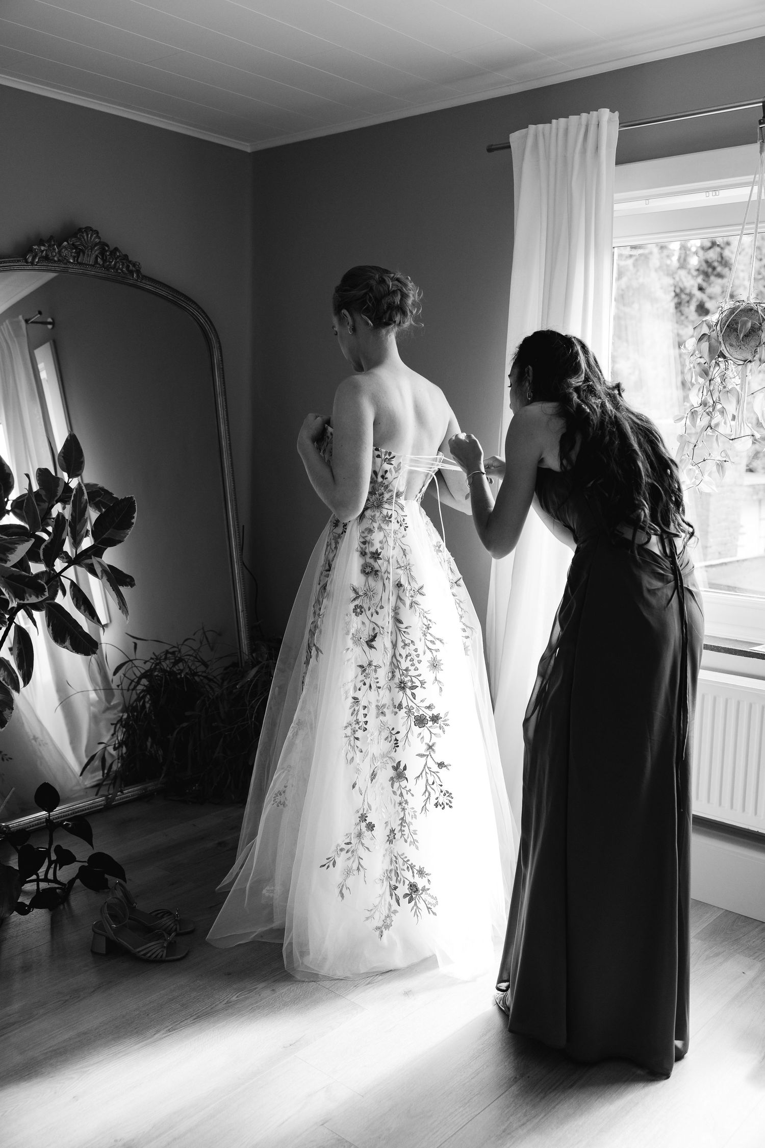 Black and white photo of bridesmaid helping bride into floral wedding dress near ornate mirror.