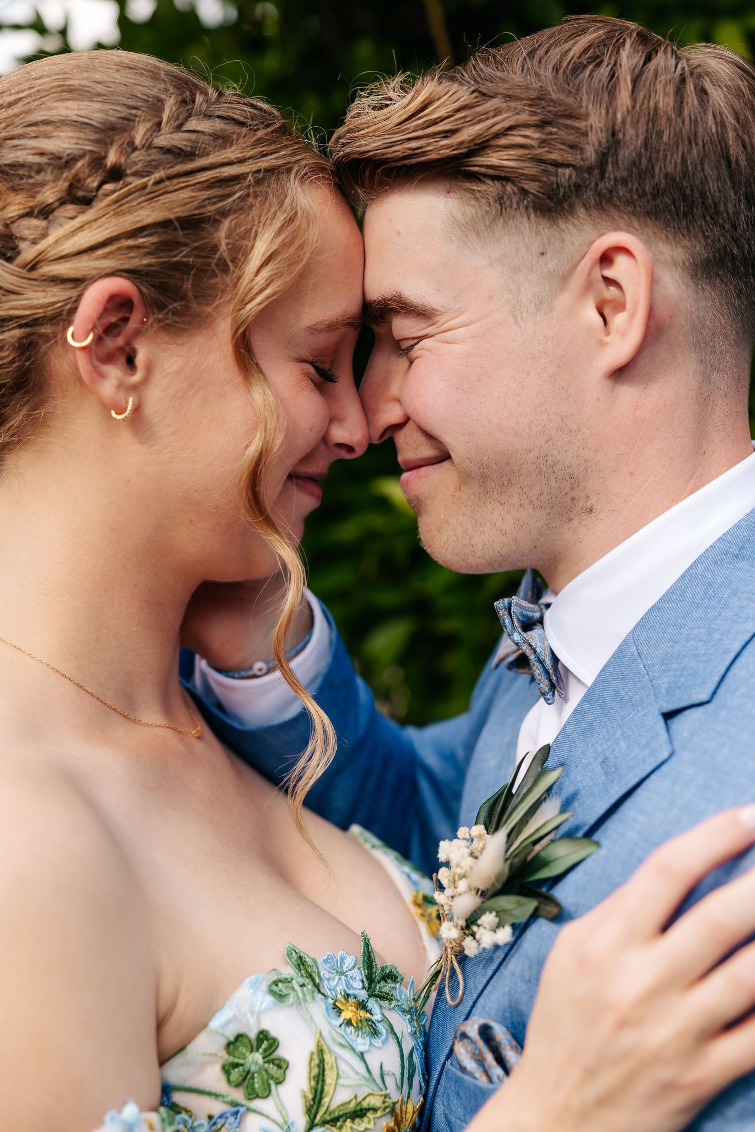 Wedding couple touches foreheads intimately, bride in floral gown and groom in light blue suit with boutonniere.