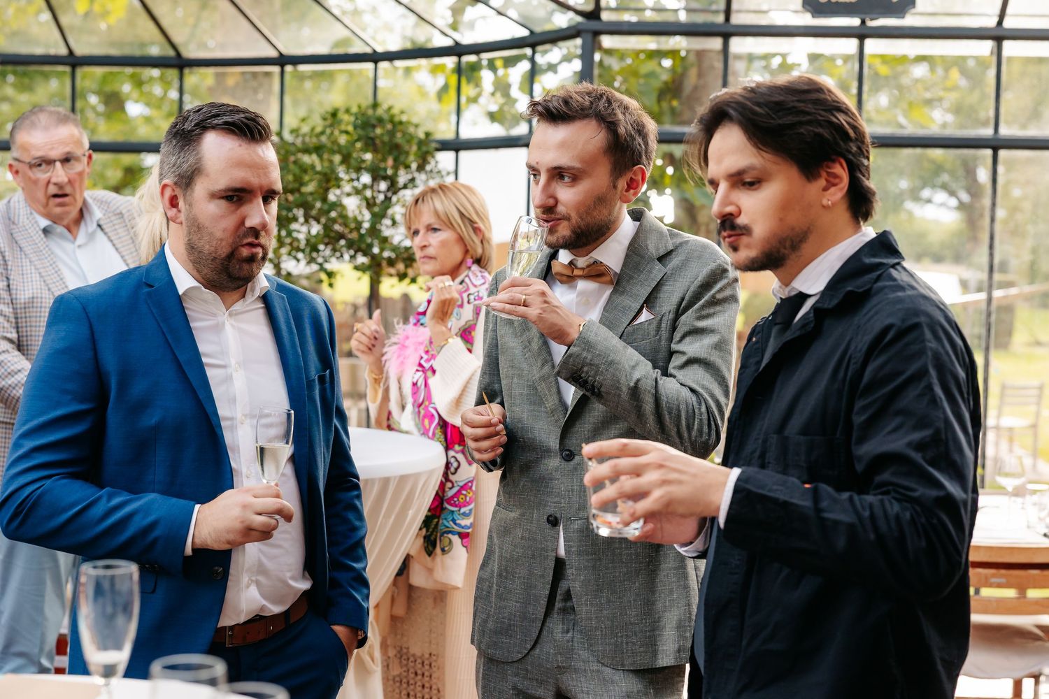 Three male wedding guests in suits conversing and holding drinks at an outdoor wedding reception.
