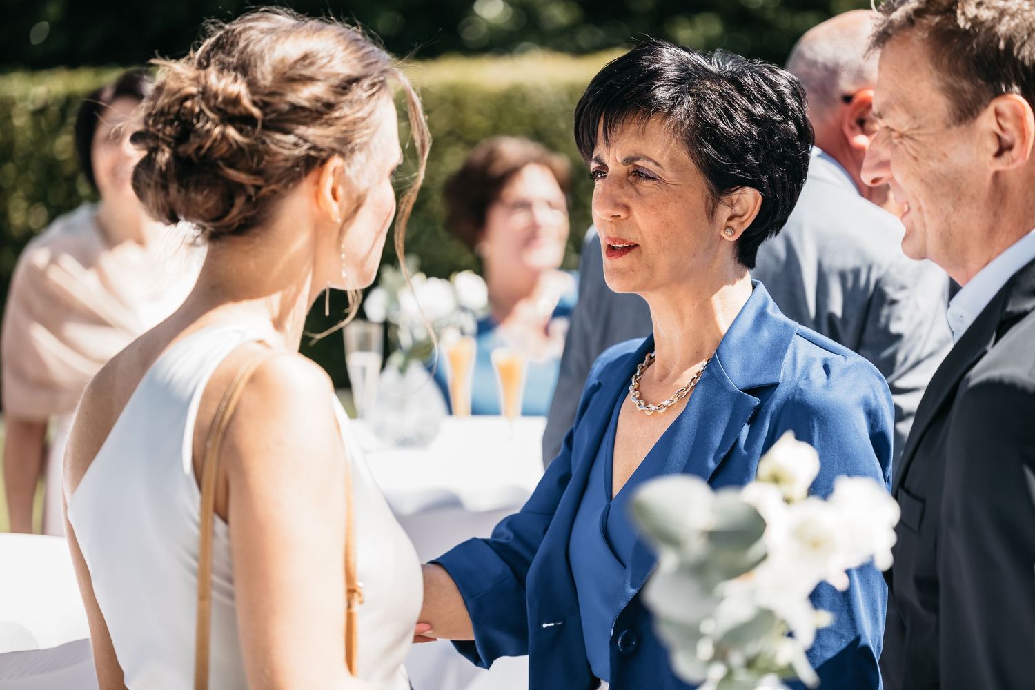 Wedding guest in blue jacket shaking hands with bride in white dress at outdoor ceremony.