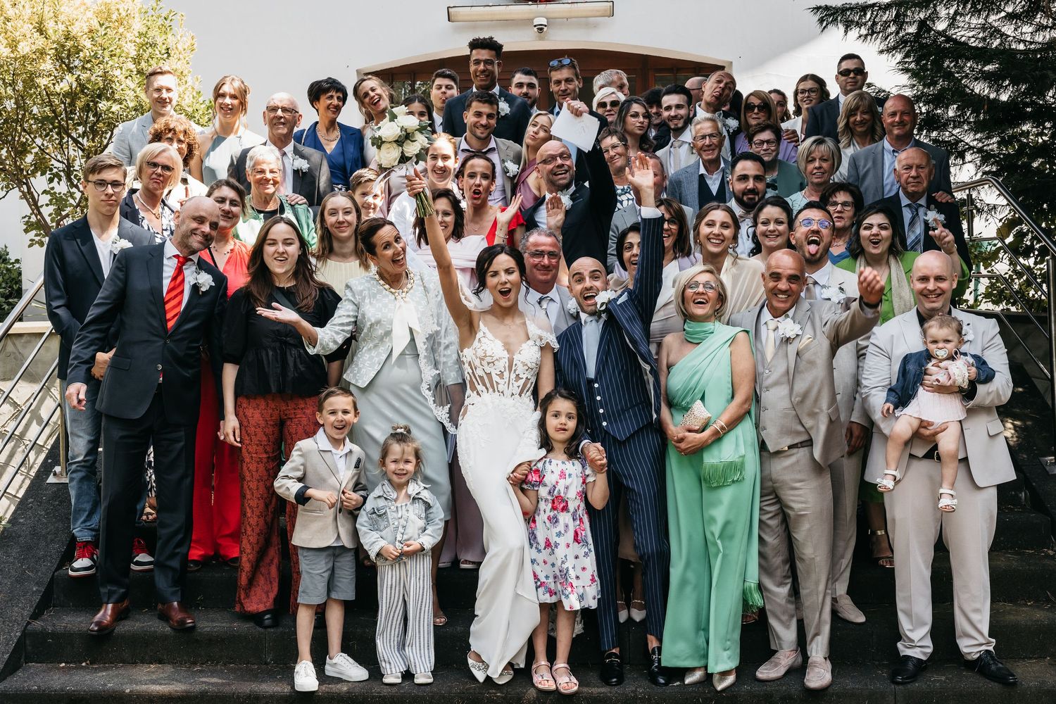 Large wedding group photo with bride and groom surrounded by smiling family and friends on steps.