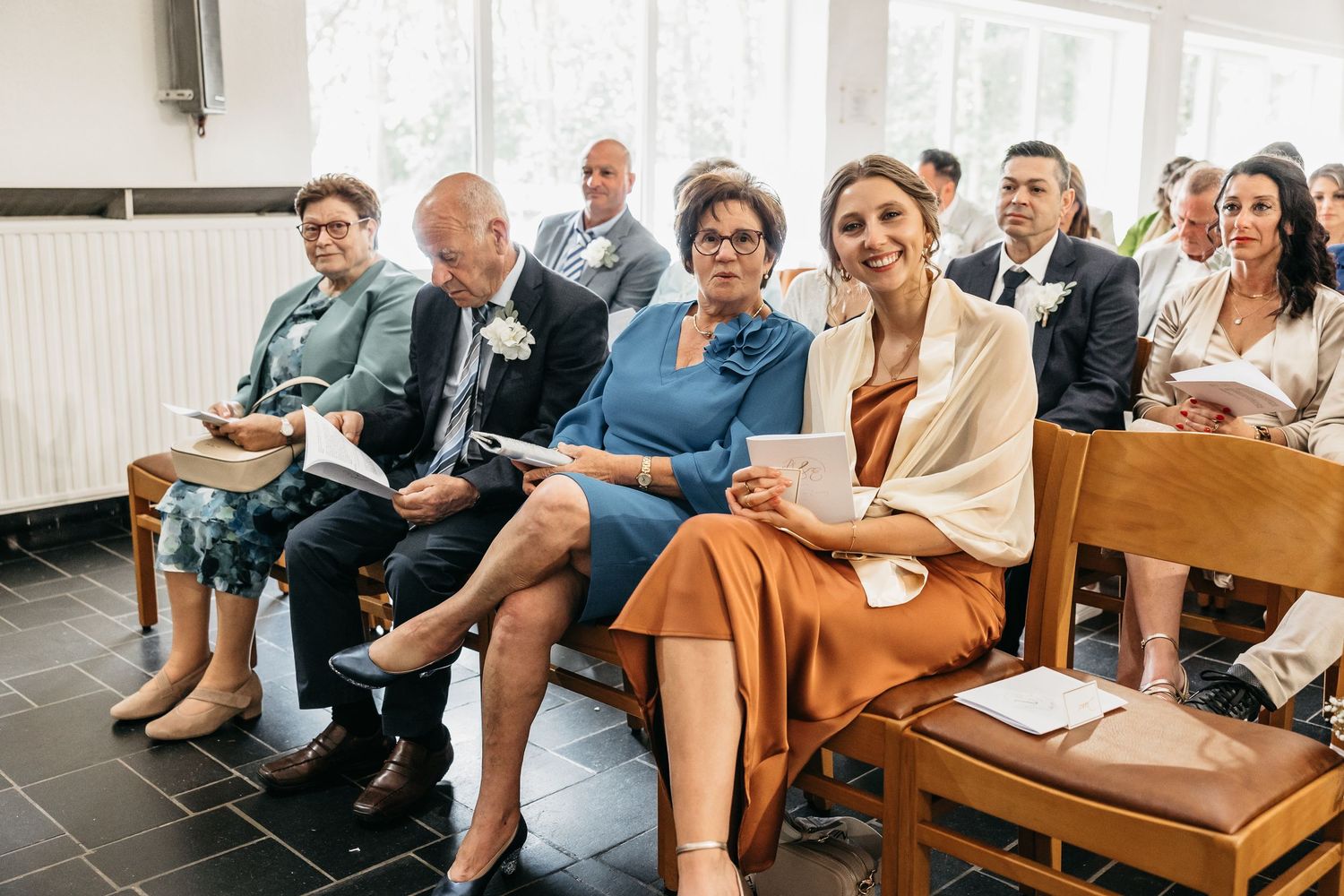 Elegantly dressed wedding guests smiling and seated on wooden church benches during a wedding ceremony.