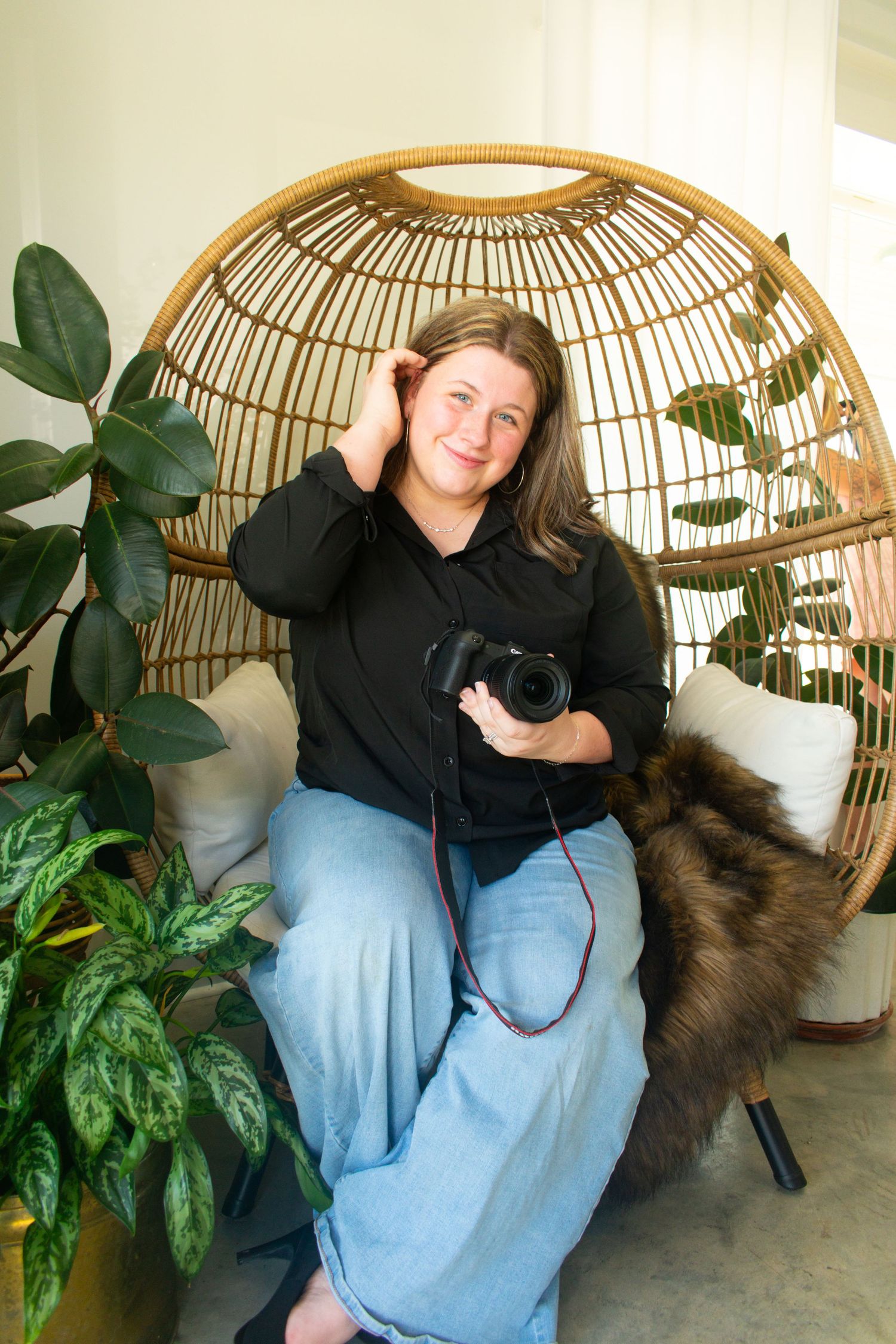 Female photographer smiling in a wicker egg chair holding a DSLR camera, surrounded by lush green indoor plants.