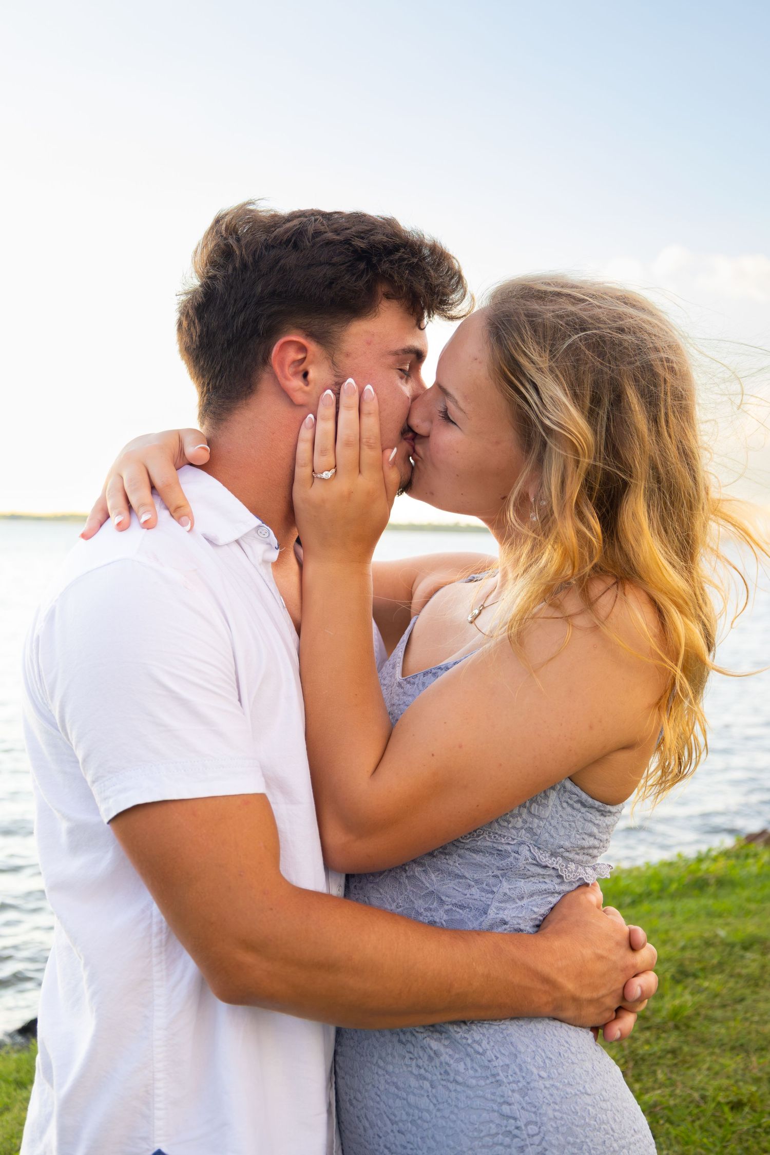 Couple sharing a romantic kiss outdoors near a lake, woman showing engagement ring, wearing casual summer outfits.