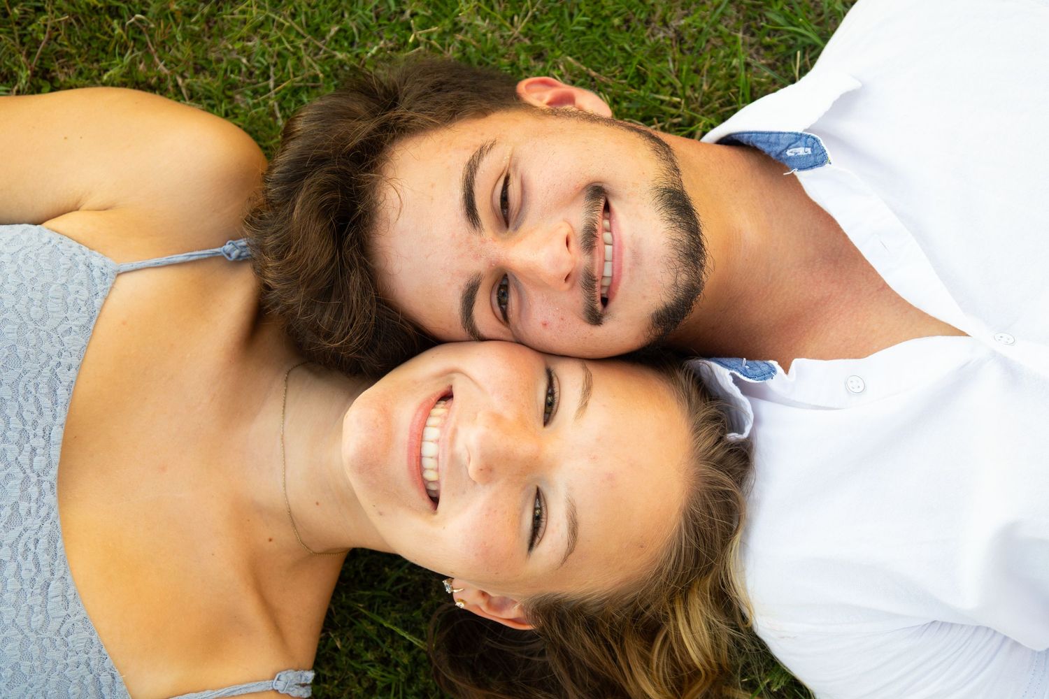 Happy young couple lying on green grass, smiling and posing head-to-head in a romantic outdoor setting.